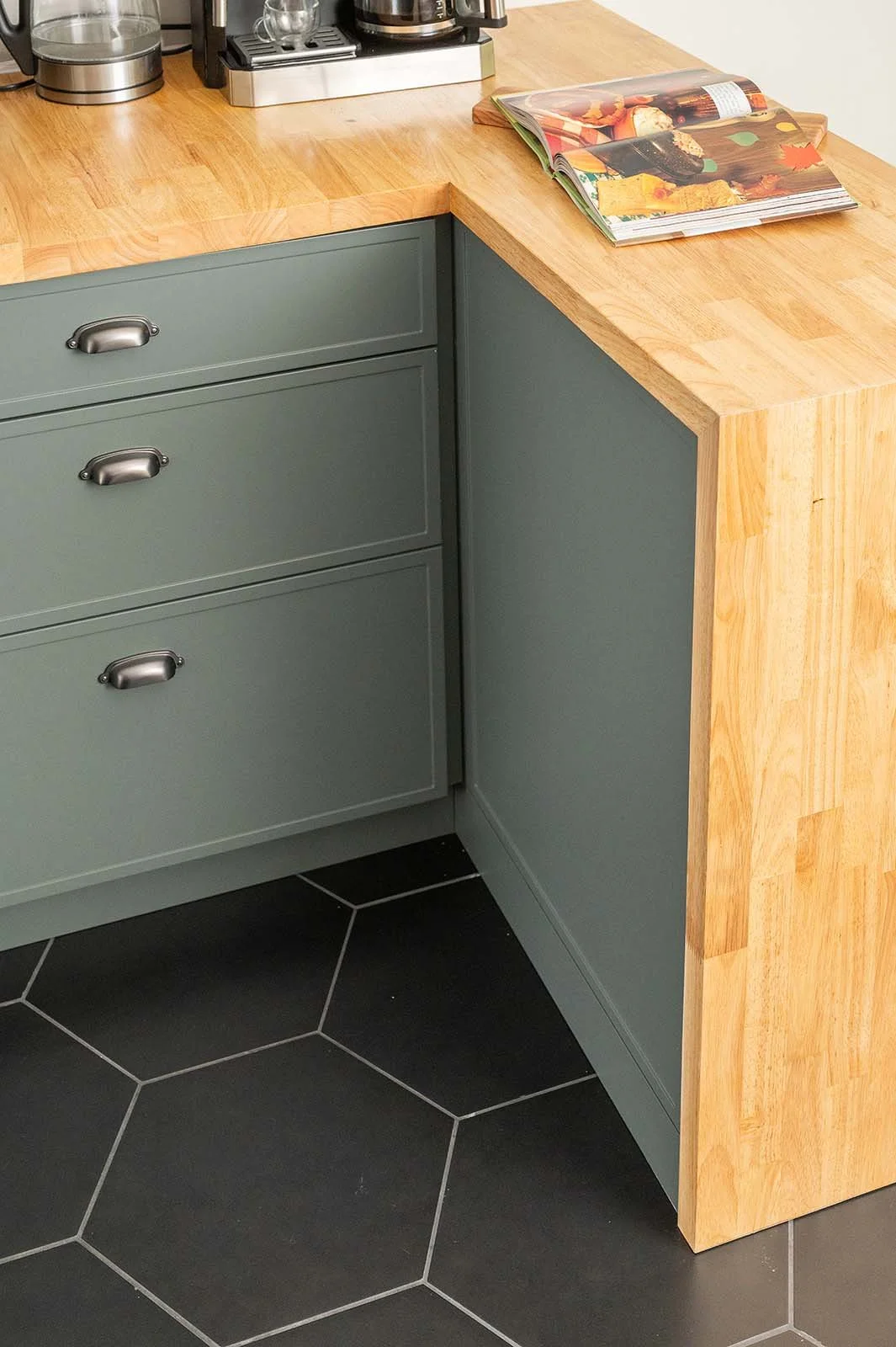 Corner of a kitchen with a wooden countertop, green cabinets with metal handles, a black tiled floor and an open magazine on the countertop.