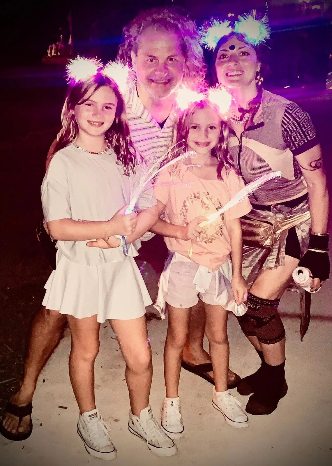 Four people, two children and two adults, celebrating at night with sparklers and glowing headbands, likely at a fireworks event.