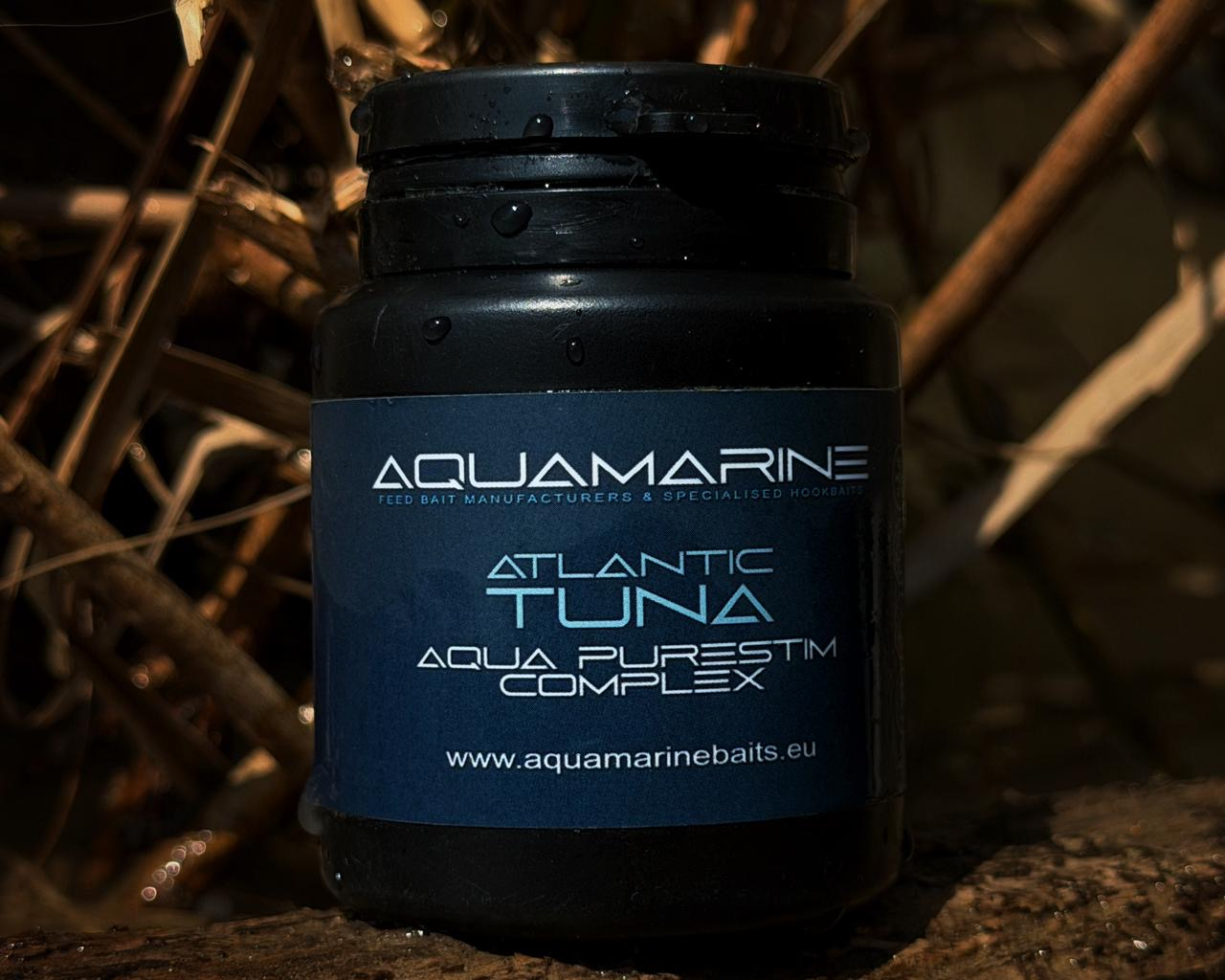 A black container of Aquamarine Atlantic Tuna Aqua Purest™ Complex fish bait on a wooden surface, with a background of dried plants or reeds.