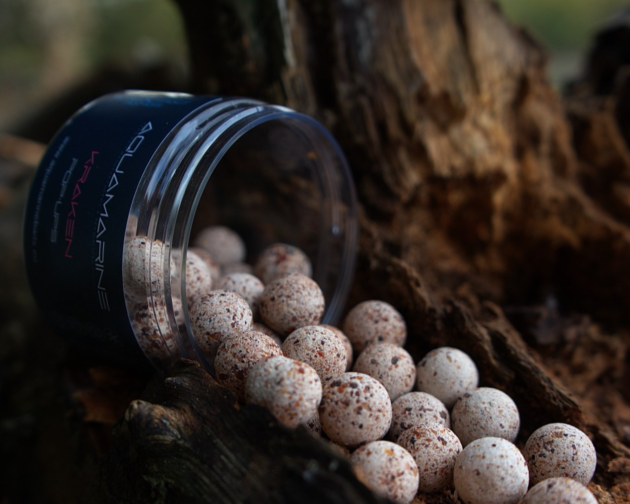 A container labeled 'Aquarium Larvae' spilled on a log with speckled white and brown pearls outside in a natural setting.