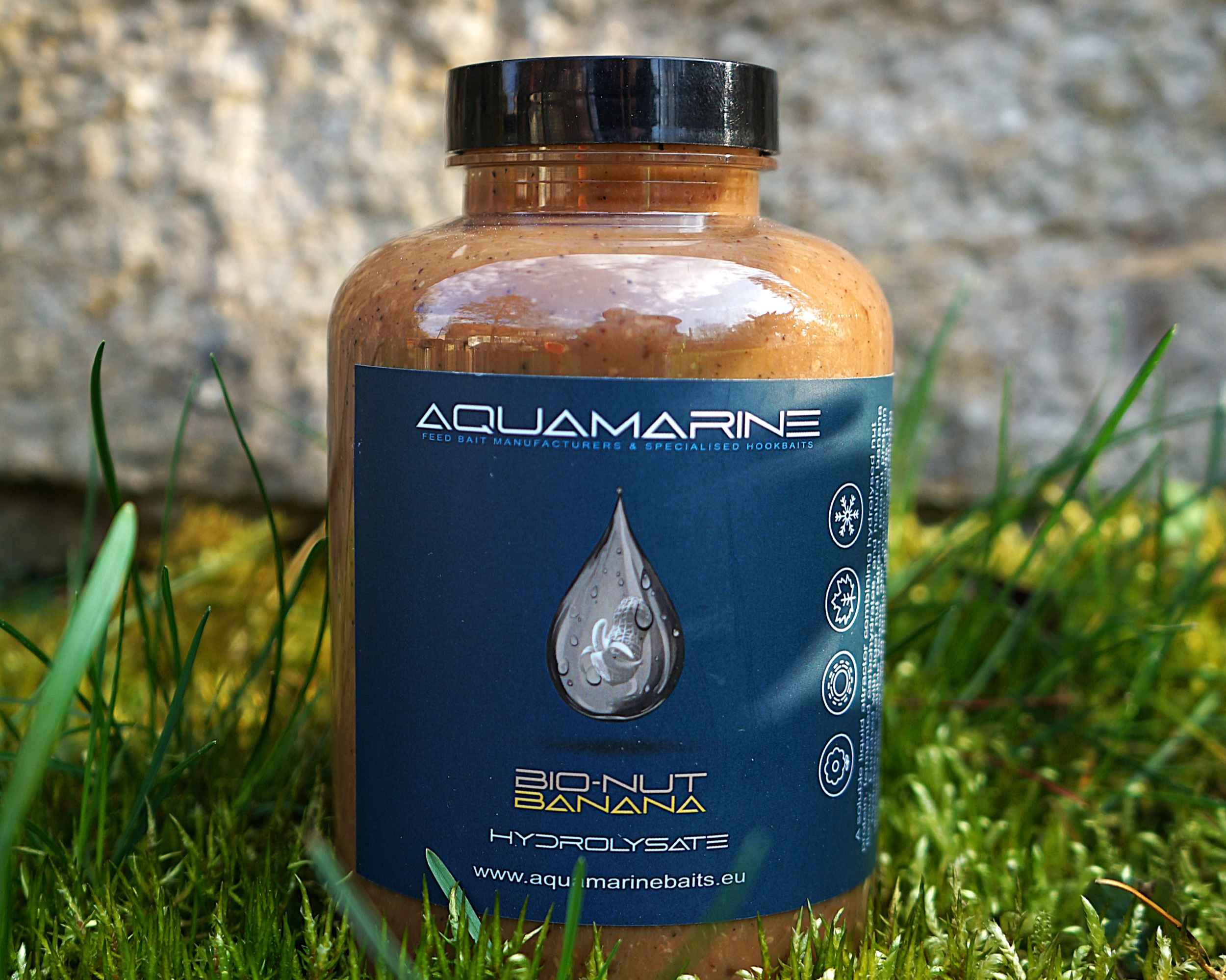 A bottle of Aquamarine Bio-Nut Banana hydrolysate bait placed on grass with a stone background.