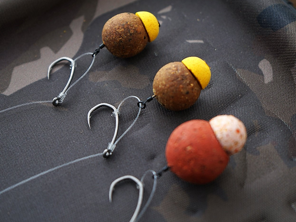 Fishing hooks with weighted bait, including two brown and yellow bait balls and one reddish-brown bait ball with white top, on a dark fabric surface suitable for fishing.