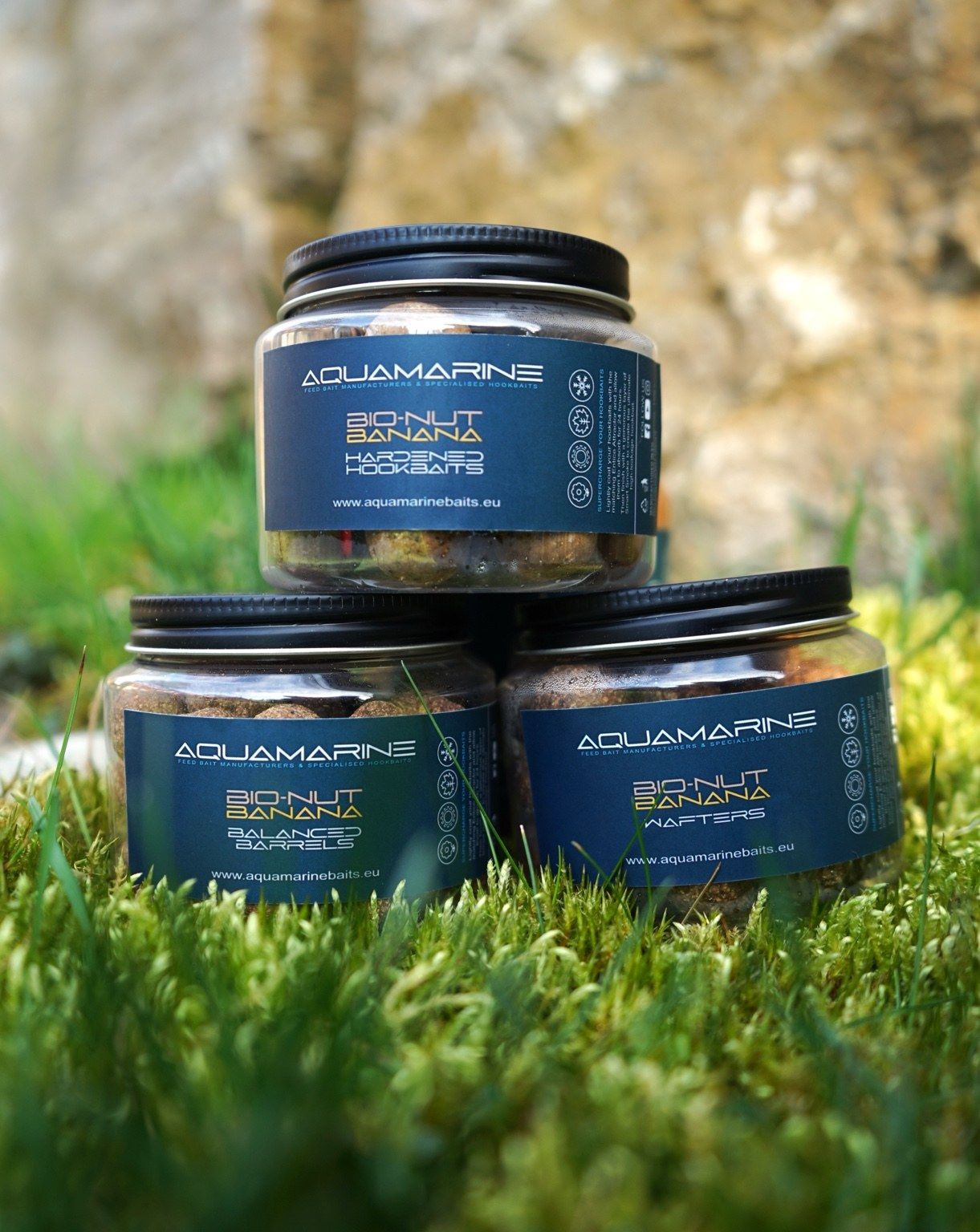 Three jars of Aquamarine Bio-Nut Banana hookbaits placed on grass outdoors, with a blurred natural background.