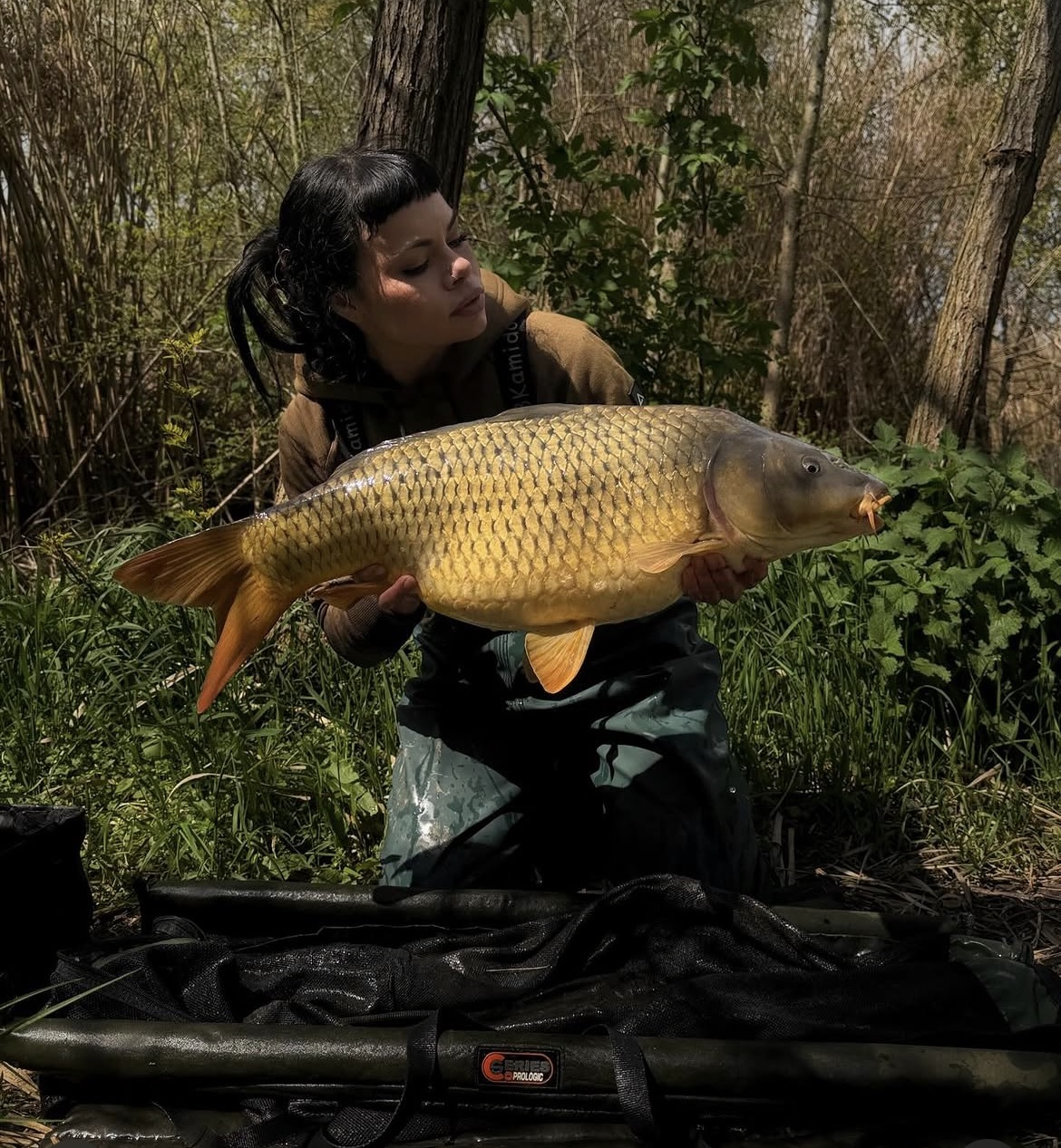 A woman holding a large carp fish in a forested area.