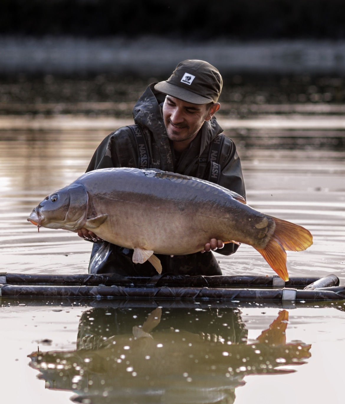 A man wading in a lake holding a large fish he caught, with a happy expression, during sunset or sunrise.