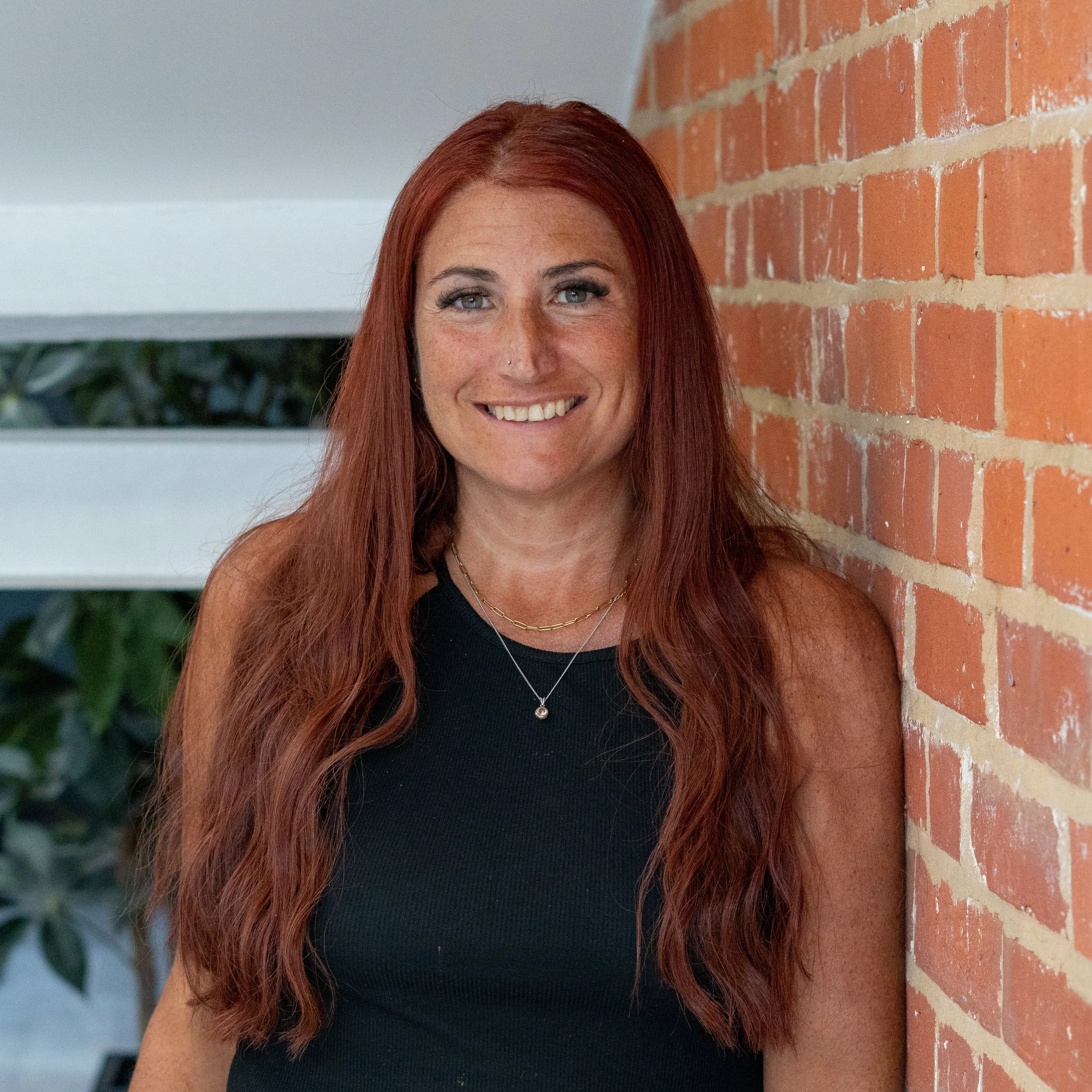 A woman with shoulder-length brown hair, smiling, wearing a pink top with black buttons, leaning against a brick wall in an office setting.