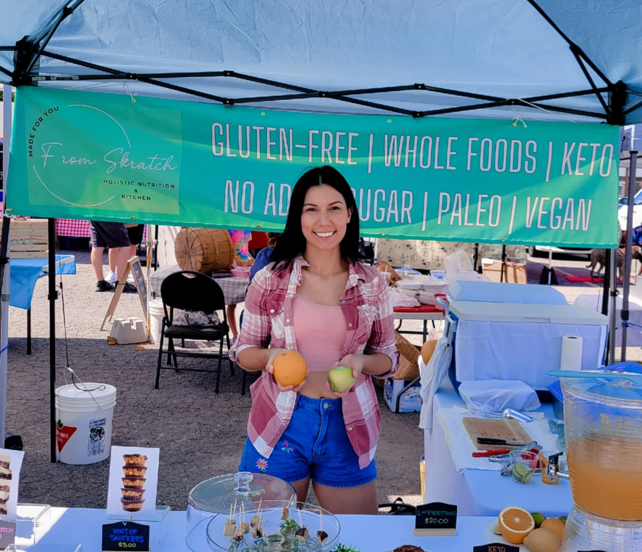 Woman at a market booth labeled "Gluten-Free, Whole Foods, Keto, Paleo, Vegan" holding a mango and an apple. Display of snacks and a drink dispenser on the table.