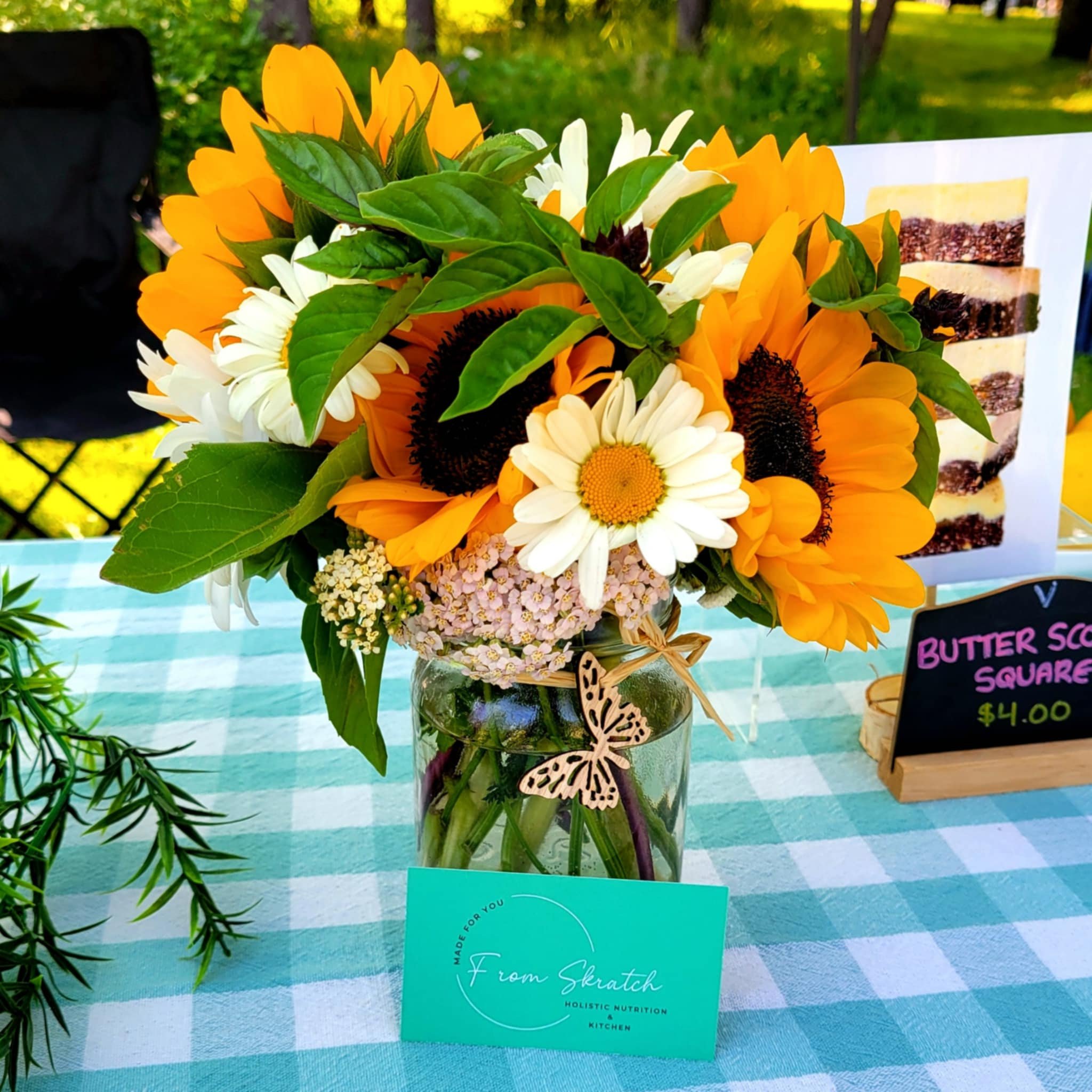 Bouquet of sunflowers, daisies, and greenery in a jar on a checkered tablecloth with a small wooden butterfly ornament. Nearby is a sign displaying 'Butter Scotch Squares $4.00' and a card labeled 'From Skratch Holistic Nutrition Kitchen.'