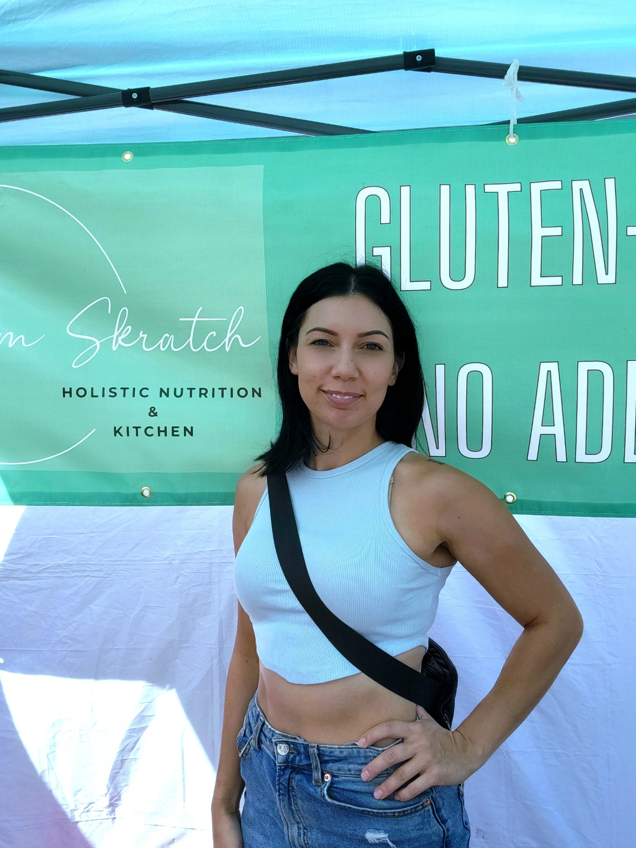 Woman in a blue crop top and denim jeans stands in front of a booth with a green banner that reads "Holistic Nutrition & Kitchen - Gluten-Free No Added Sugar."