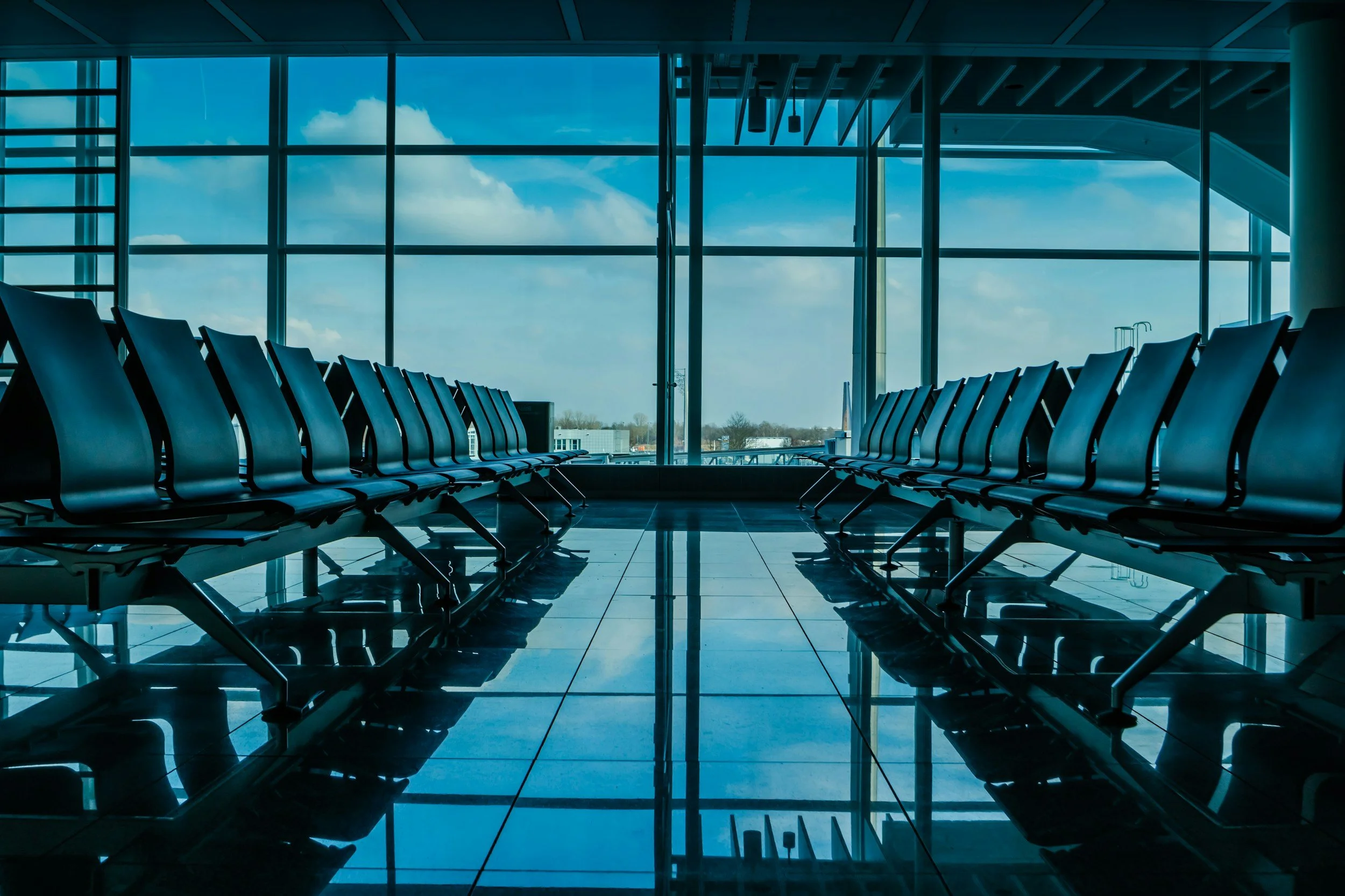 Empty airport waiting area with black seats, large windows, and a blue sky with clouds outside.