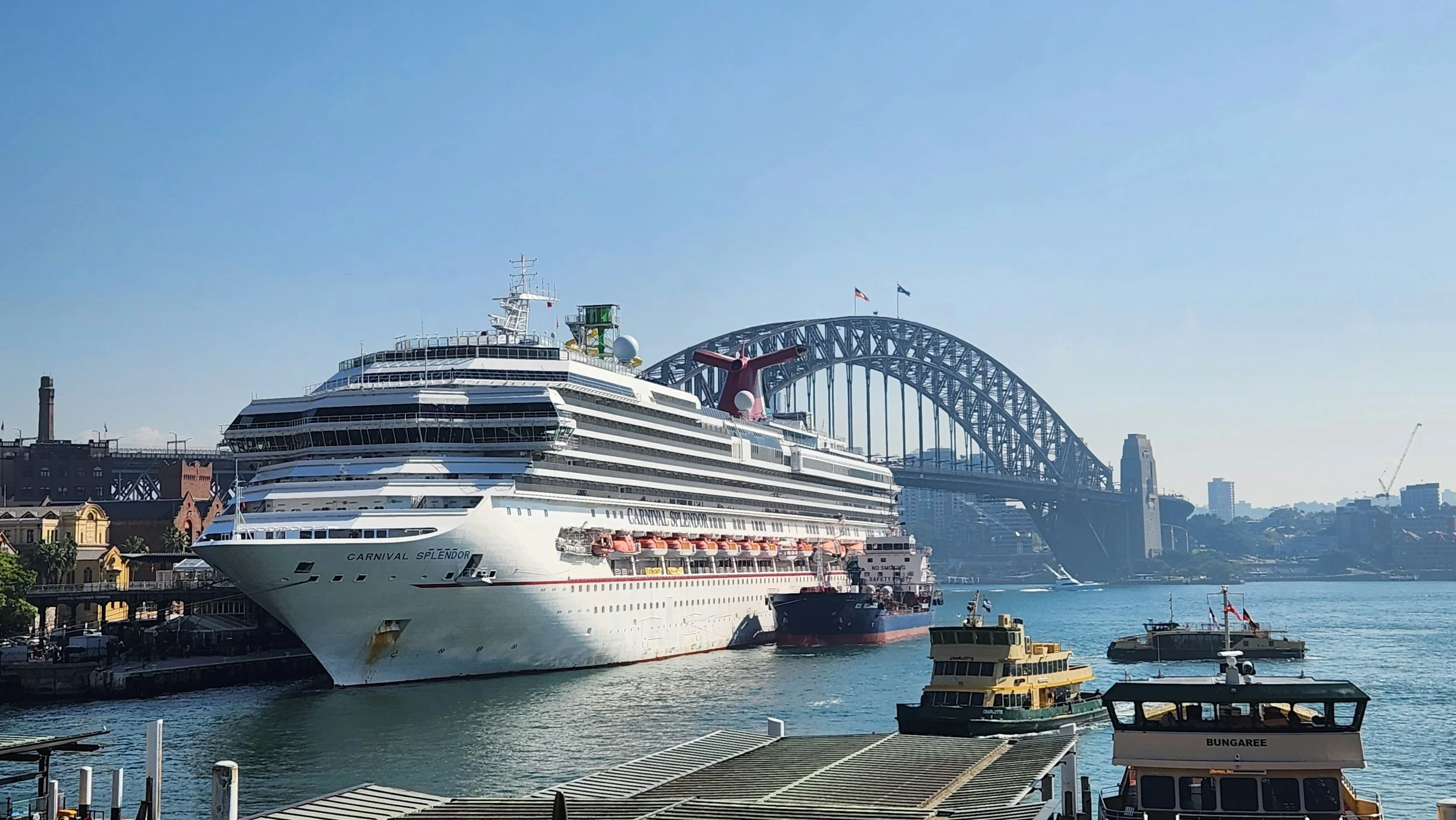A large cruise ship named Carnival Splendor docked at a harbor with smaller boats in the water and a bridge in the background on a clear day.