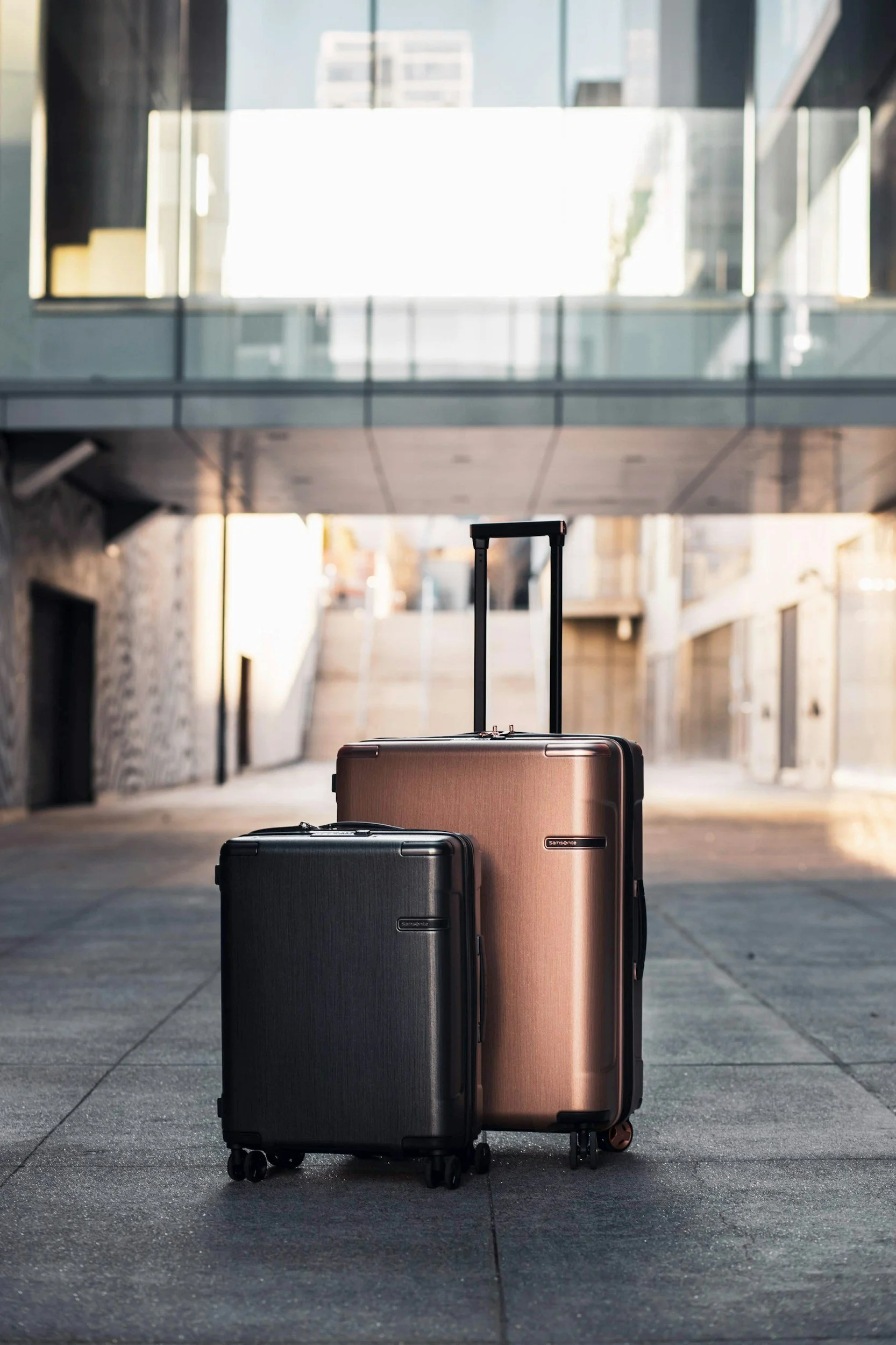 Two suitcases, one black and one rose gold, standing on an urban sidewalk with modern buildings and a bridge above in the background.