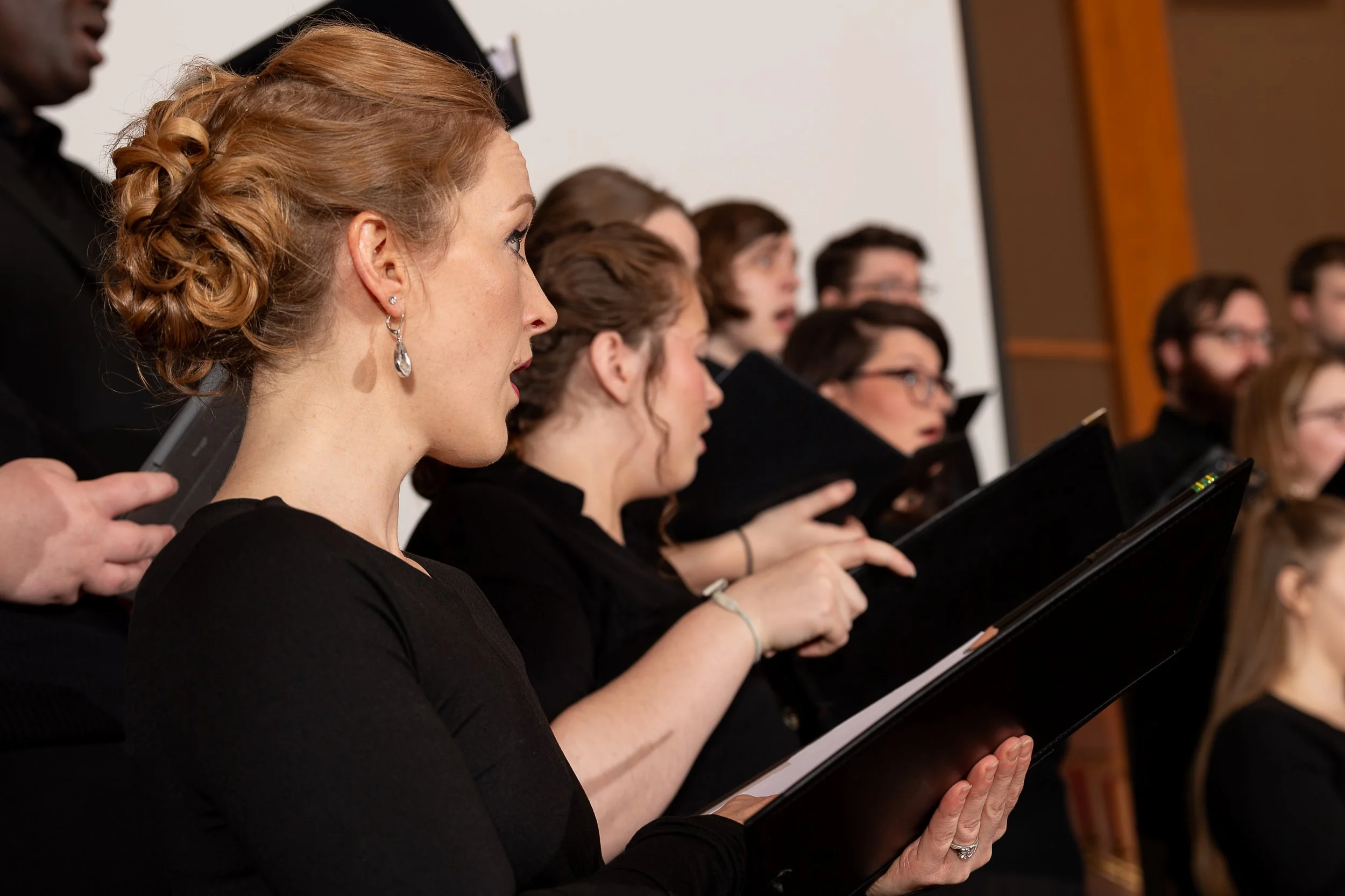 A group of choir singers holding sheet music and singing, dressed in black, with a focus on a woman with red hair and drop earrings in the foreground.