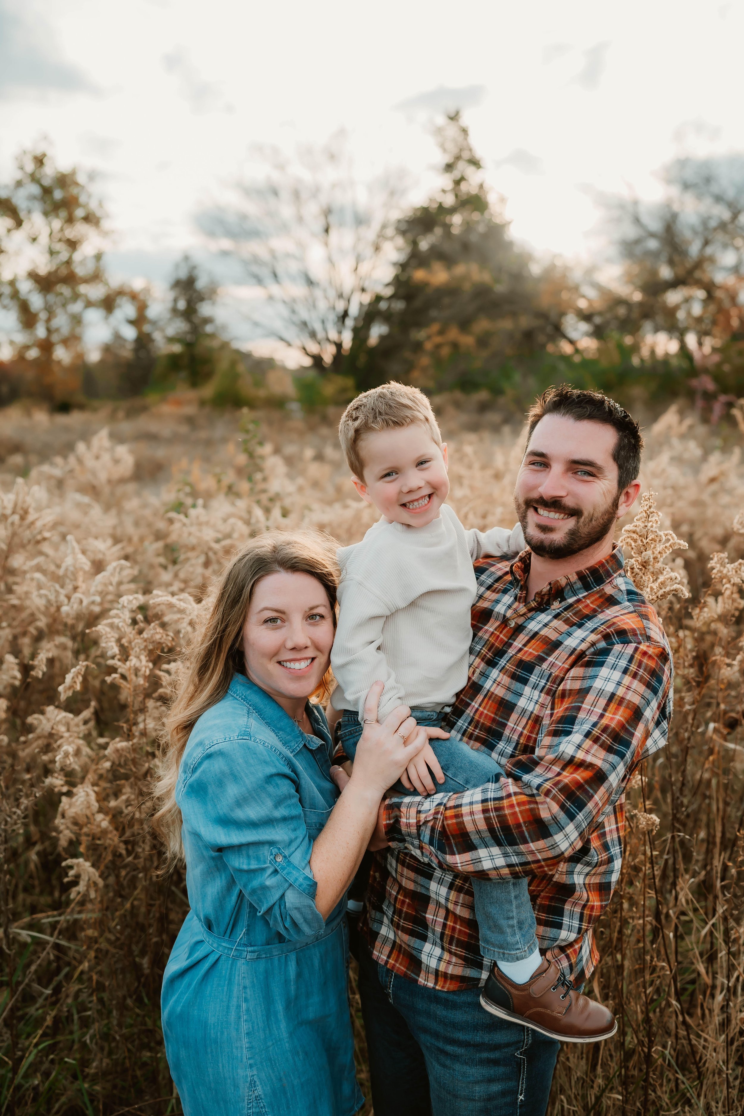 Family of three outdoors, with the man holding a smiling boy, and the woman standing beside them, in a field of tall dried grasses during sunset, with trees and a cloudy sky in the background.