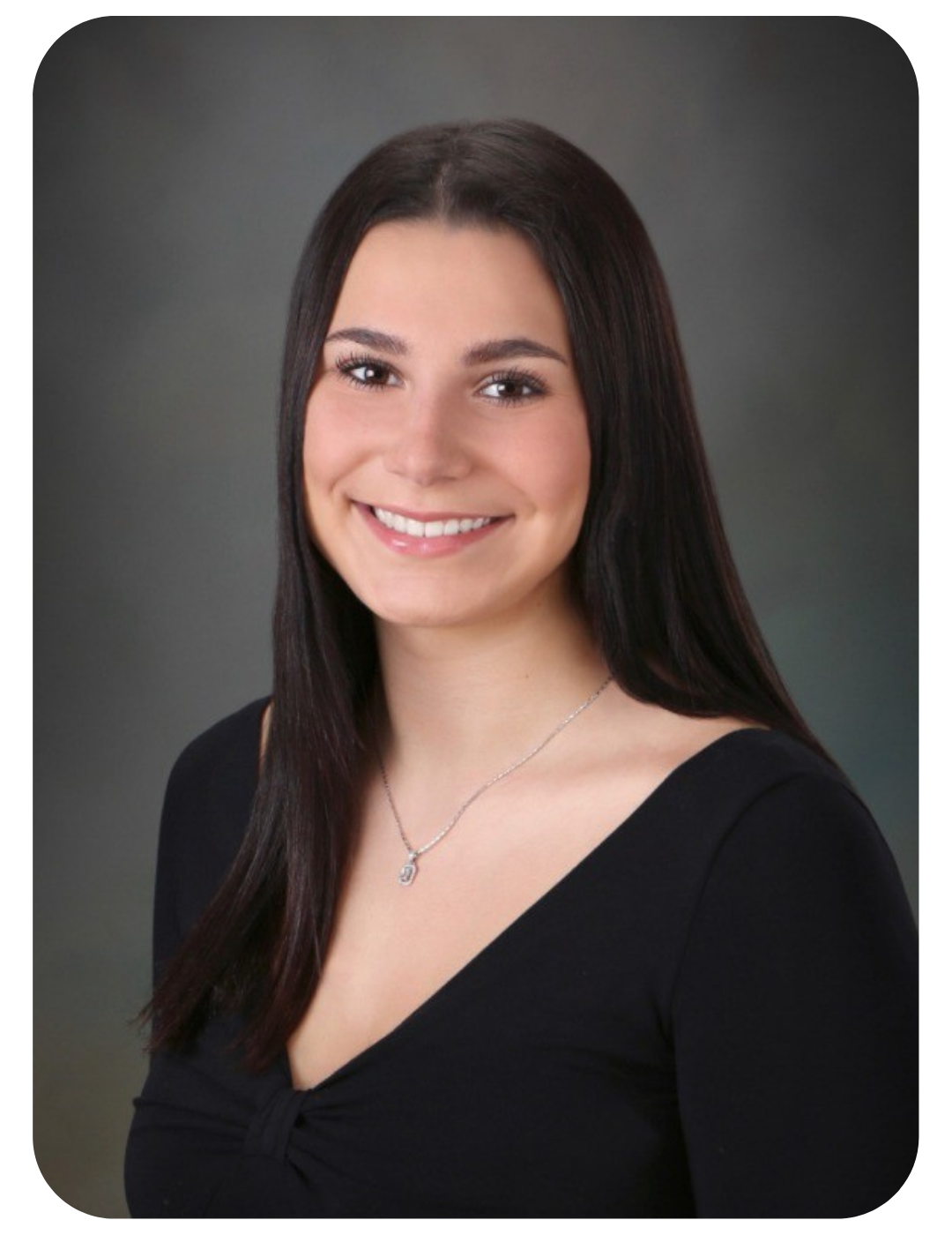 A woman with long brown hair, wearing a diamond necklace and a black v-neck shirt, smiling brightly with a neutral gray background.