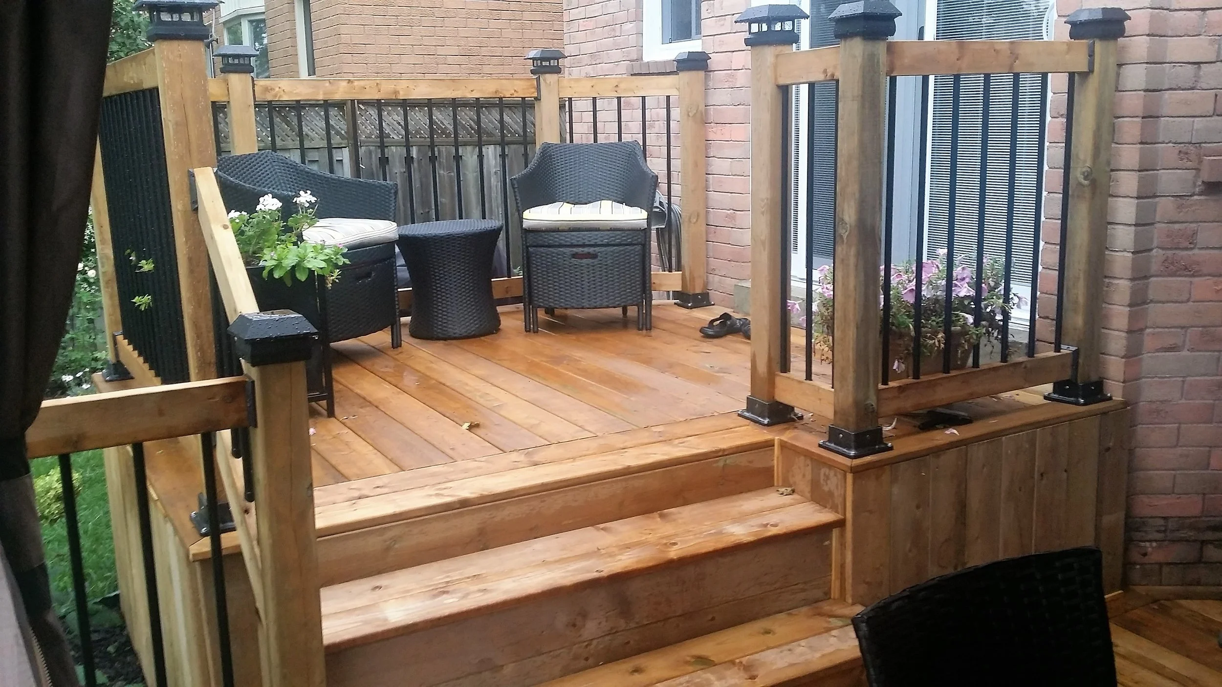 A small wooden balcony with black metal railing, two black wicker chairs with cushions, a small black wicker side table, potted plants, and a pair of sandals, attached to a brick building.