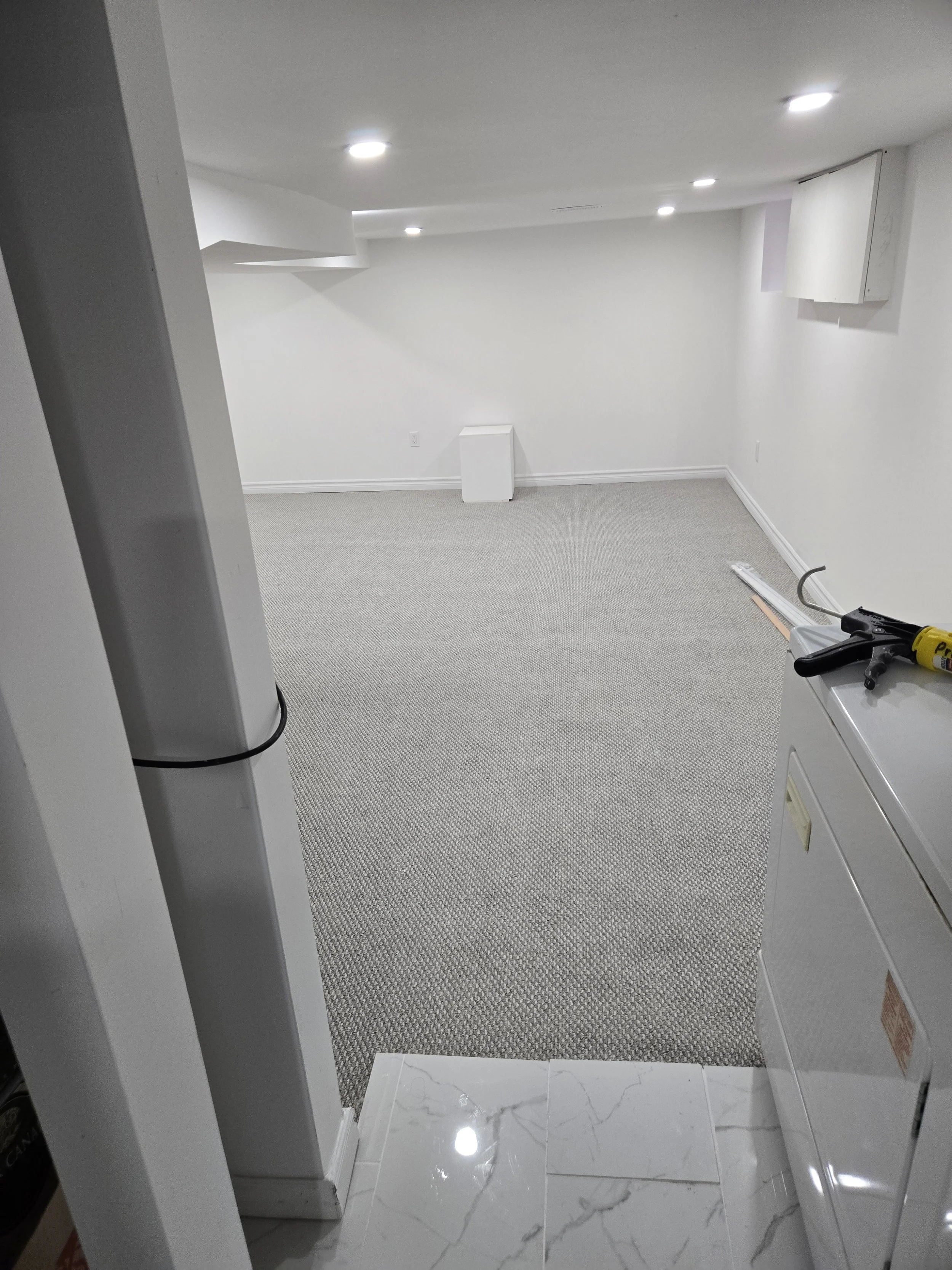 Empty white-walled basement room with white carpet, ceiling lights, and some construction tools on a nearby surface.