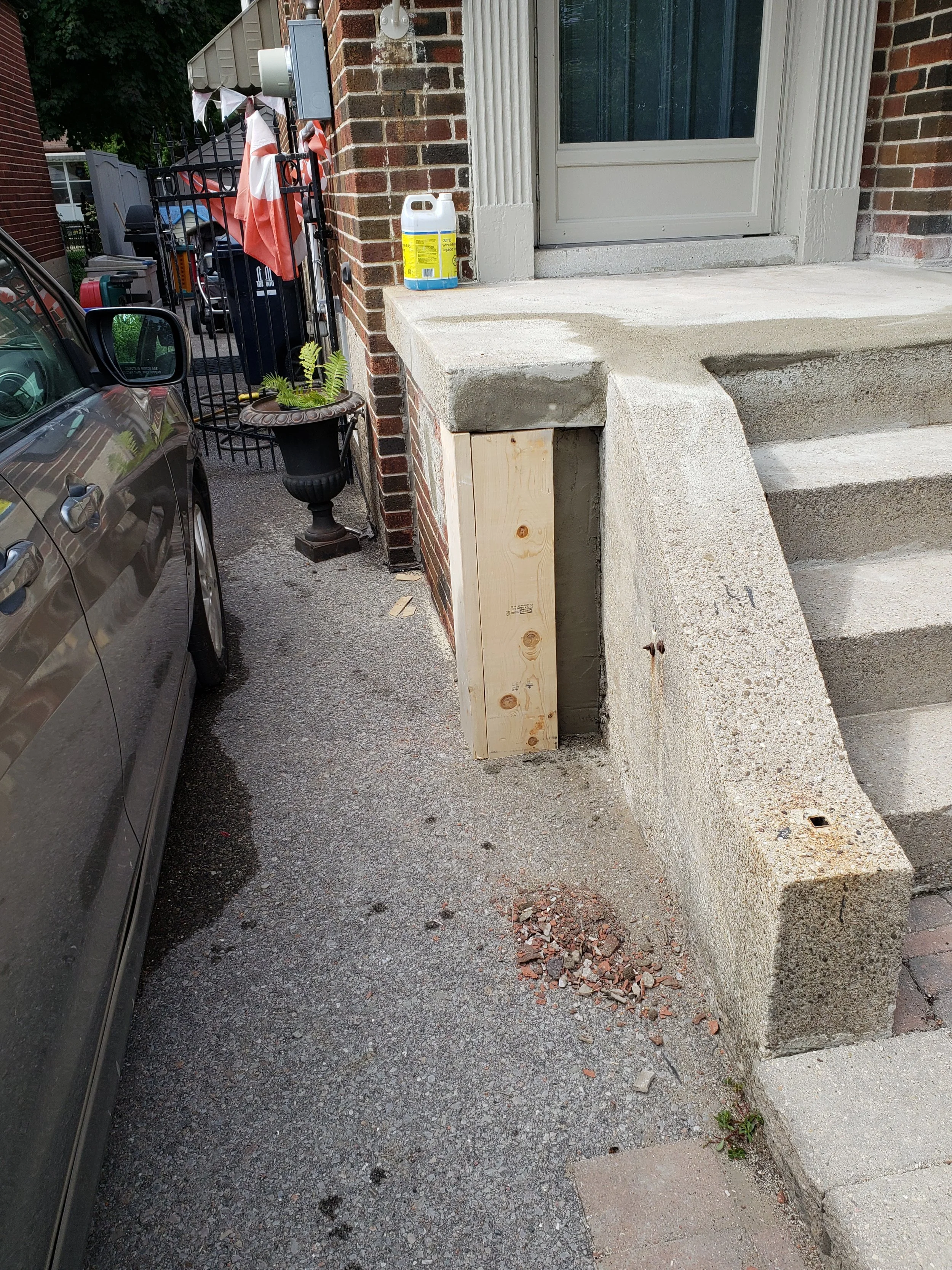 Construction of a concrete porch with a new wooden support post at the front entrance of a brick house, with a brown car parked nearby.