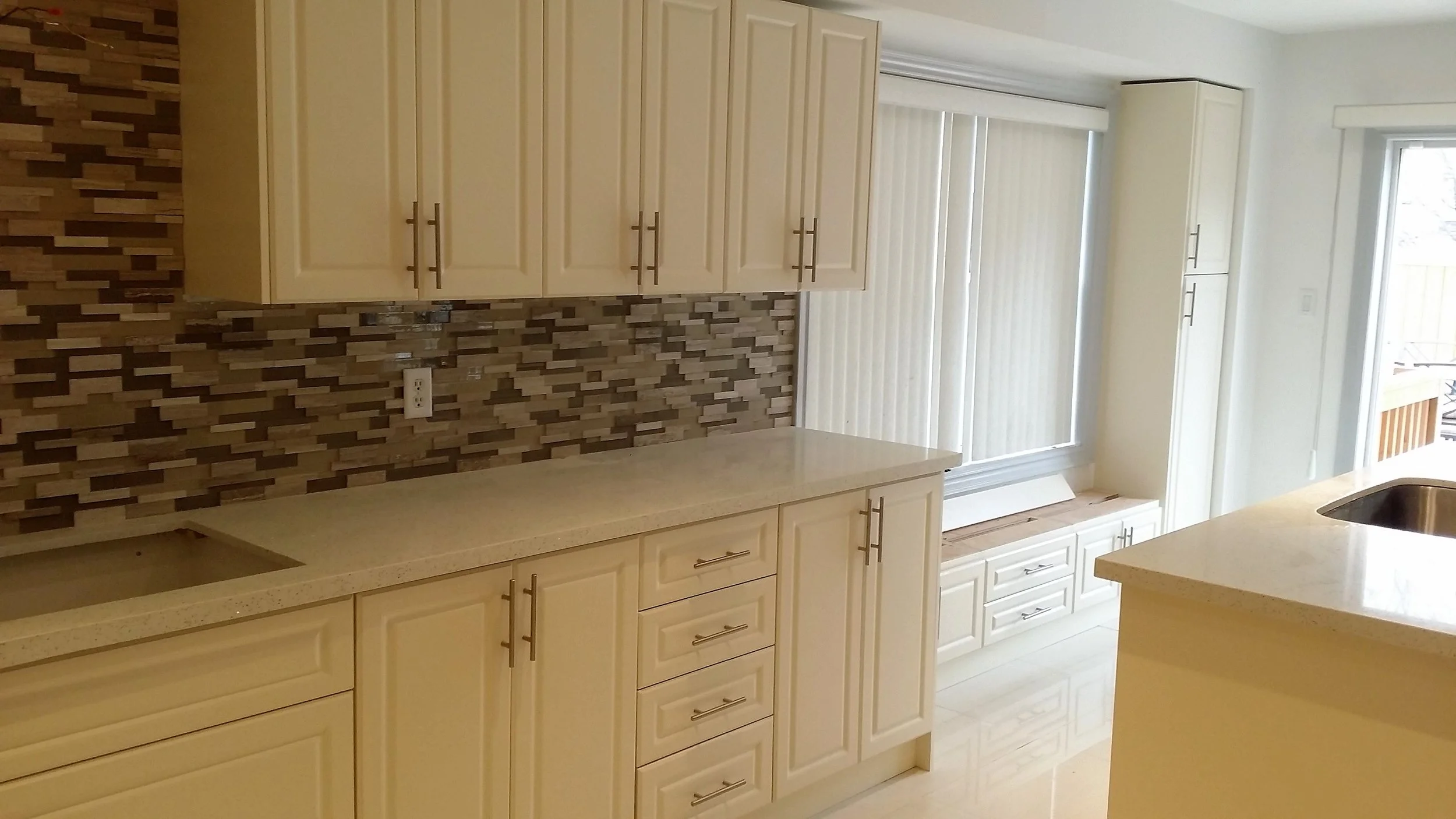 Kitchen with cream-colored cabinets, a beige countertop, and a decorative tile backsplash. There's a window with vertical blinds and a sliding glass door to an outdoor balcony.
