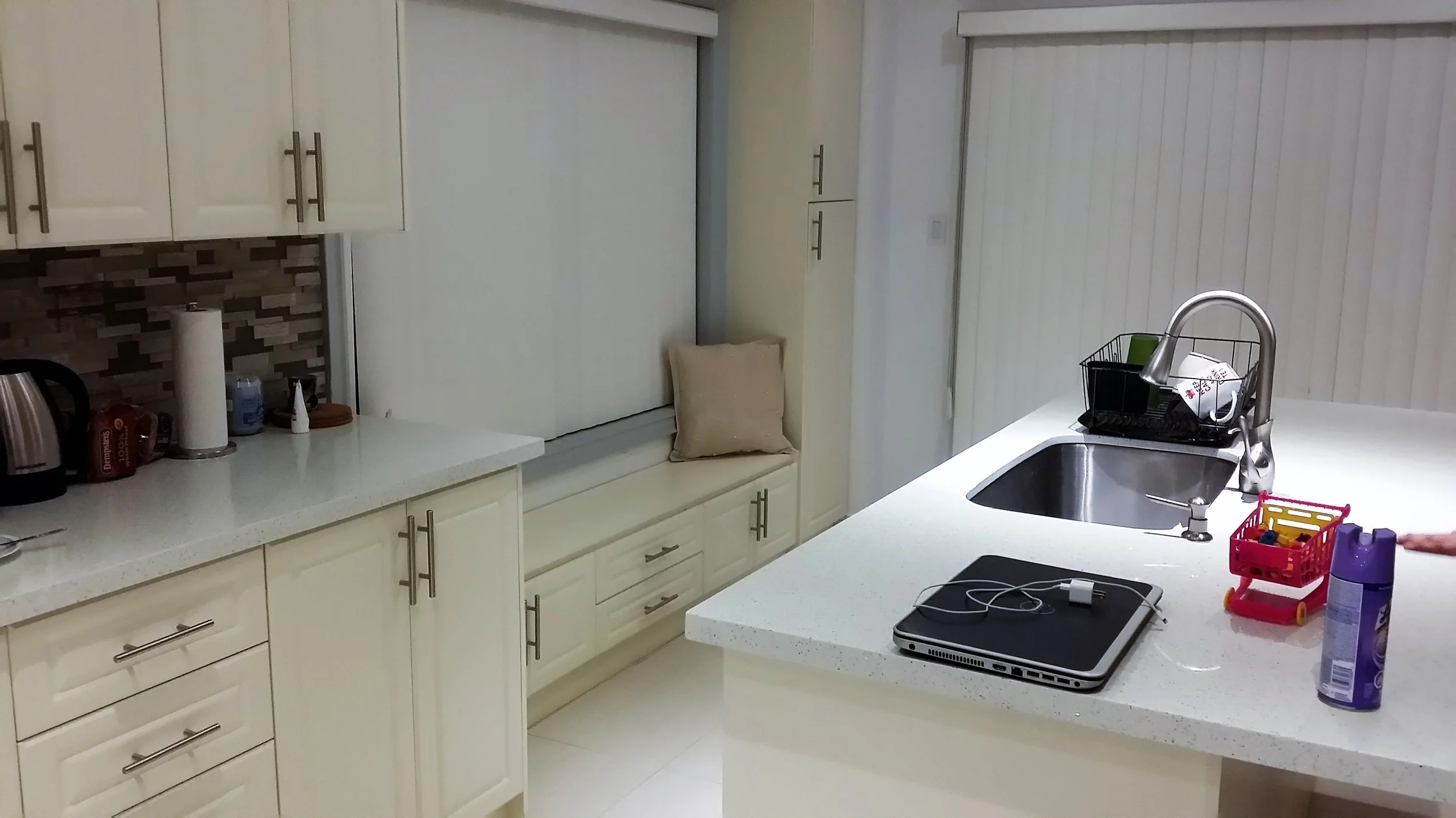 Kitchen with white cabinets, beige and brown tiled backsplash, white countertops, a sink, and various items including a laptop, phone chargers, a shopping cart, a purple bottle, and other small kitchen appliances.