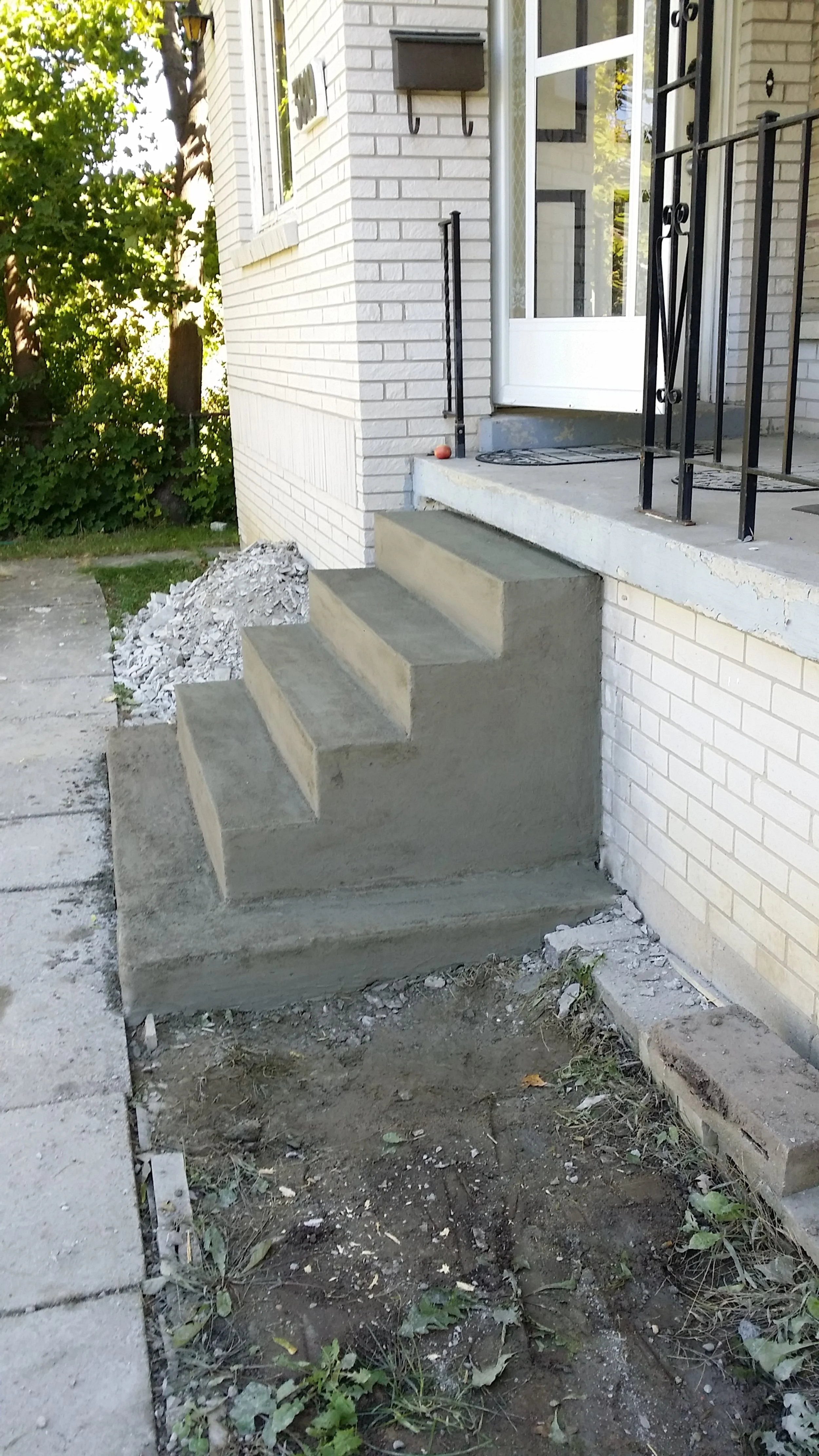 Concrete front steps under construction leading to a house with a white brick exterior, black iron railing, and front door, with construction debris and dirt nearby.