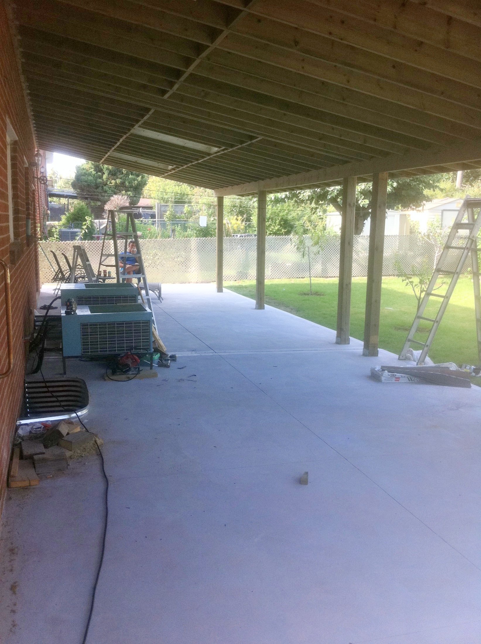 A backyard patio under construction with a concrete floor, wooden posts supporting a roof, ladders, and construction tools. There is a grassy yard, trees, and a fence in the background.