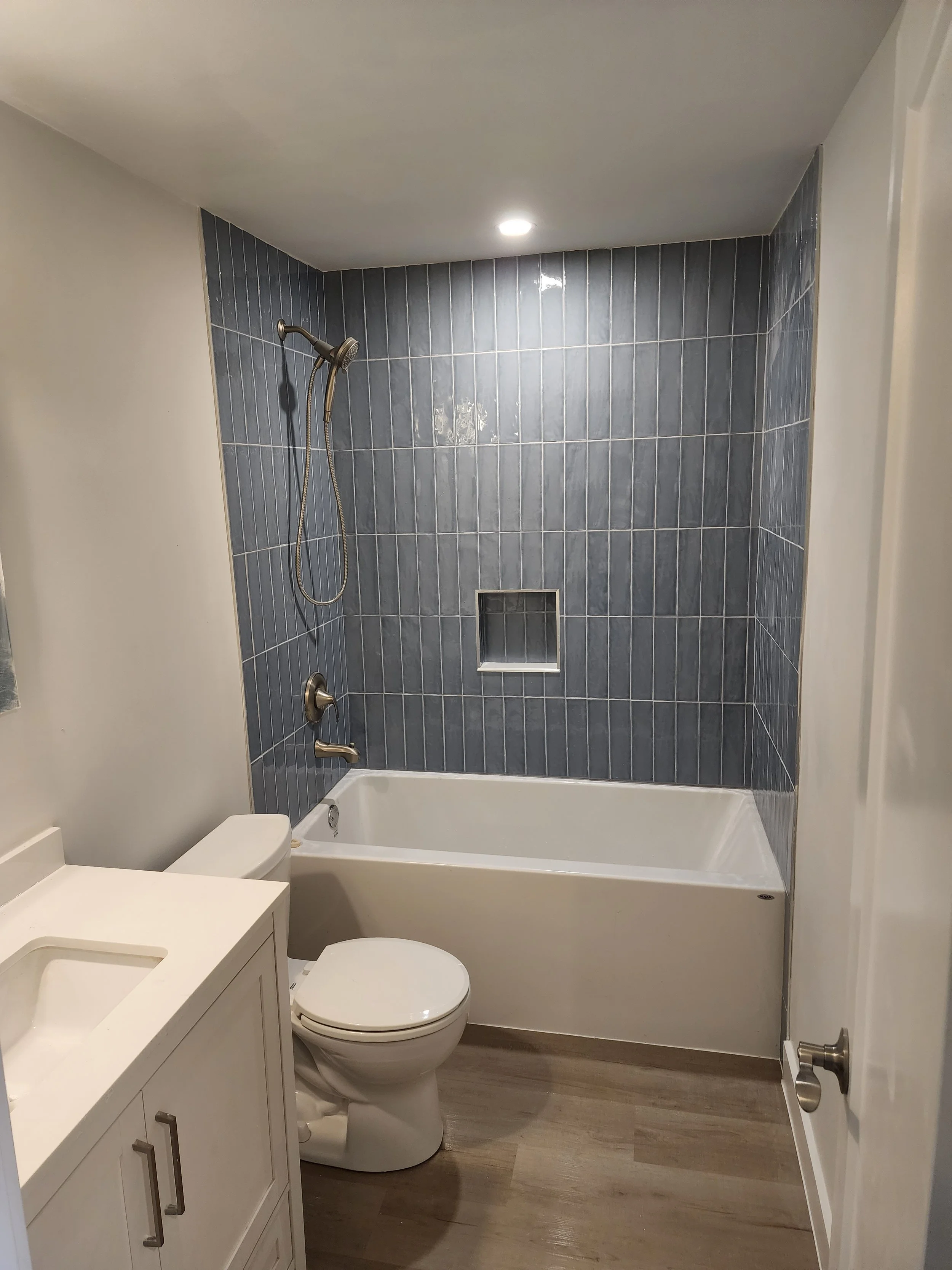 Bathroom with blue tiled wall, bathtub, toilet, small vanity with sink, and wooden flooring.