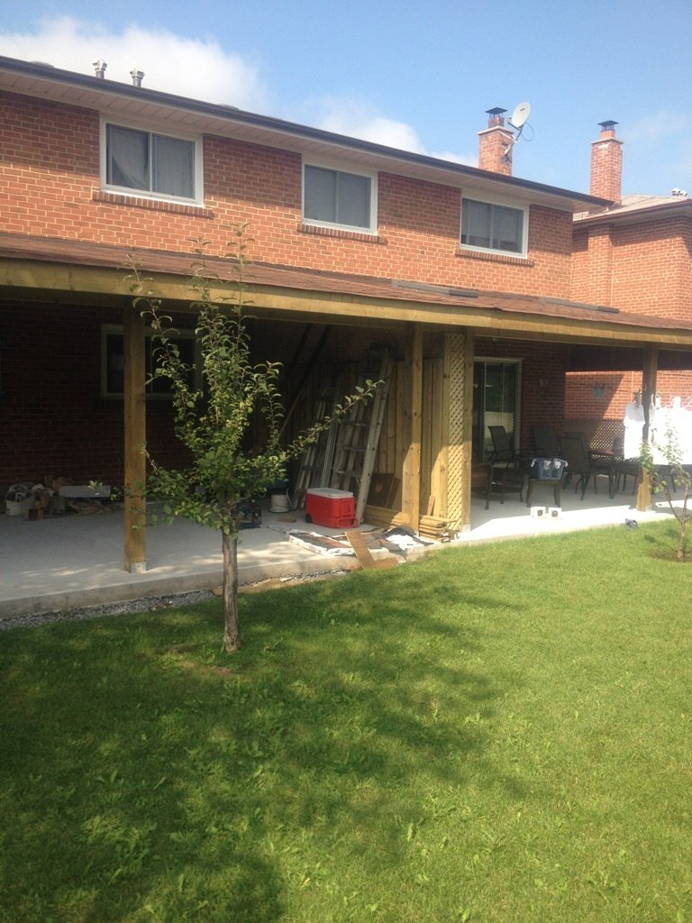 Backyard with a partially constructed wooden porch, two small trees, and outdoor furniture on a concrete patio.
