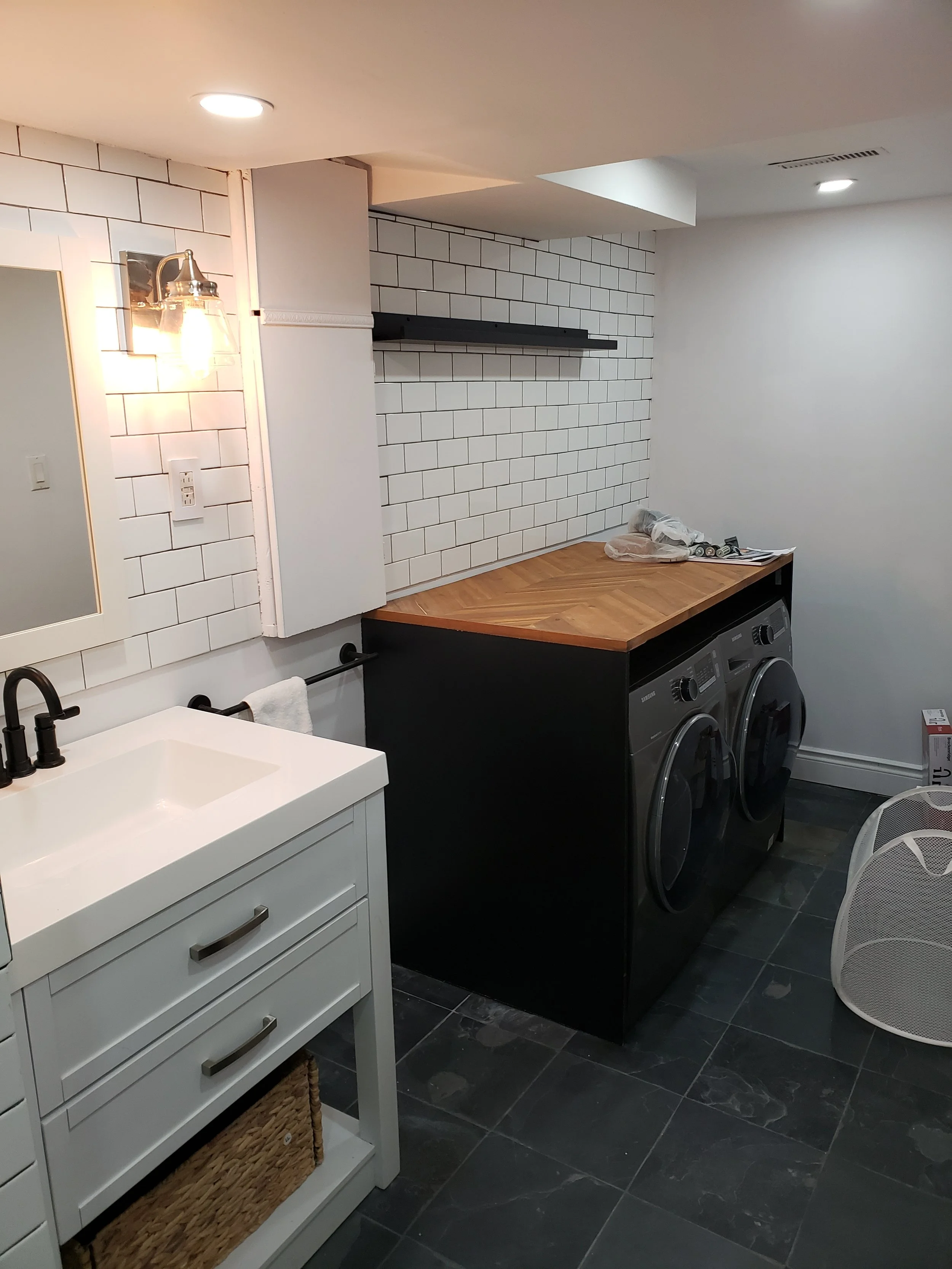 A laundry room with a white sink vanity, a mirror, black faucet, a towel bar with a towel, a black and wooden countertop, a white brick wall, and a washing machine and dryer.
