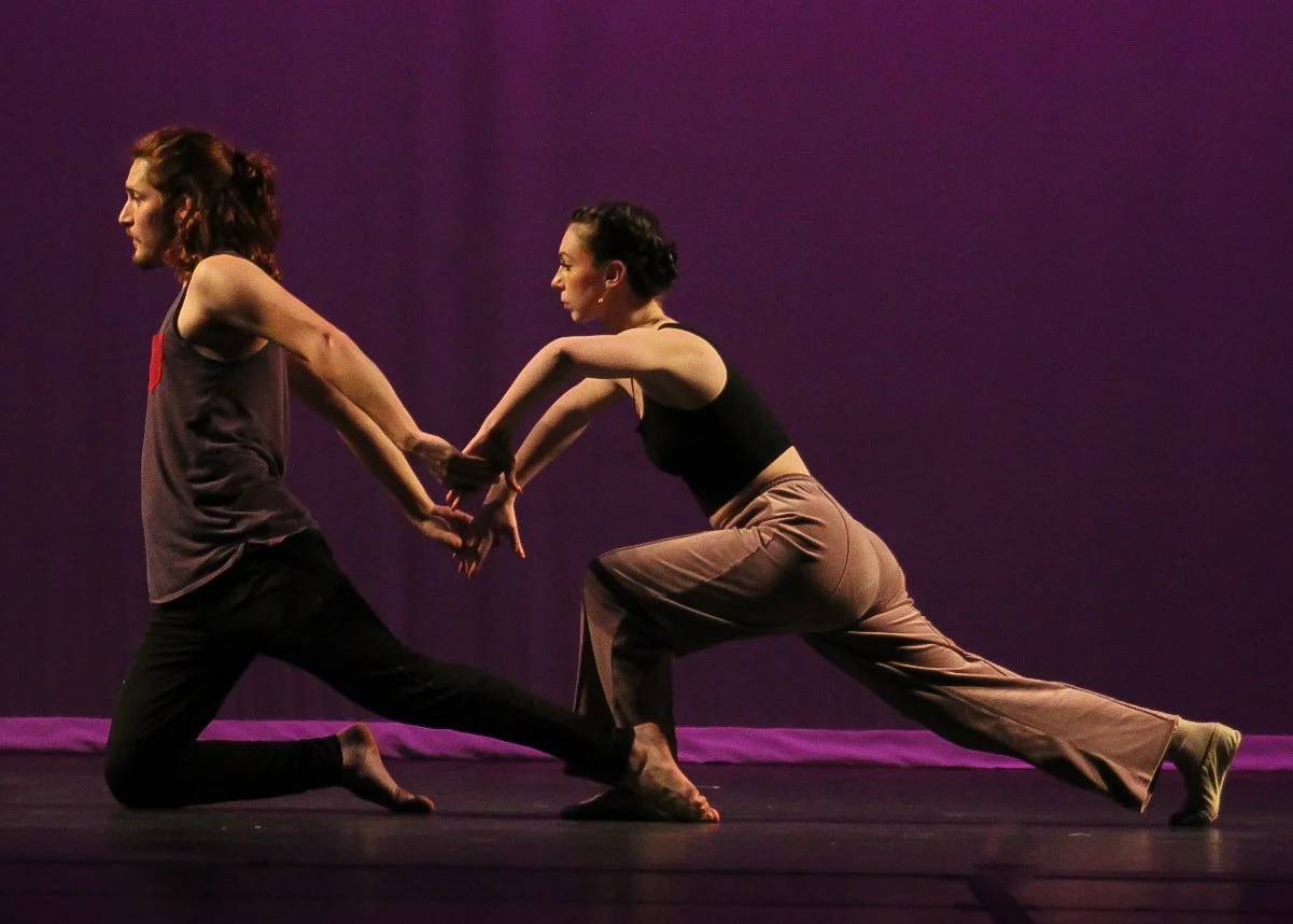 Two dancers performing on stage, holding hands. The performer on the left is kneeling on one knee, while the performer on the right is lunging forward with one leg extended behind. The stage has a purple backdrop.