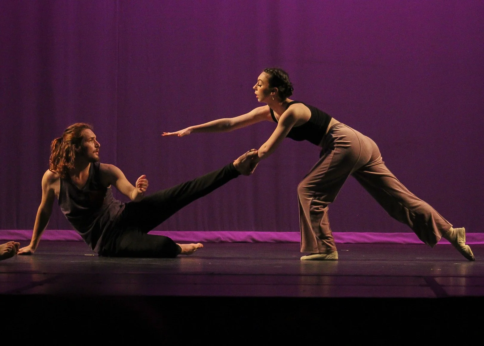 Two dancers on stage, one sitting with legs extended, the other leaning forward with arms outstretched, against a purple backdrop.