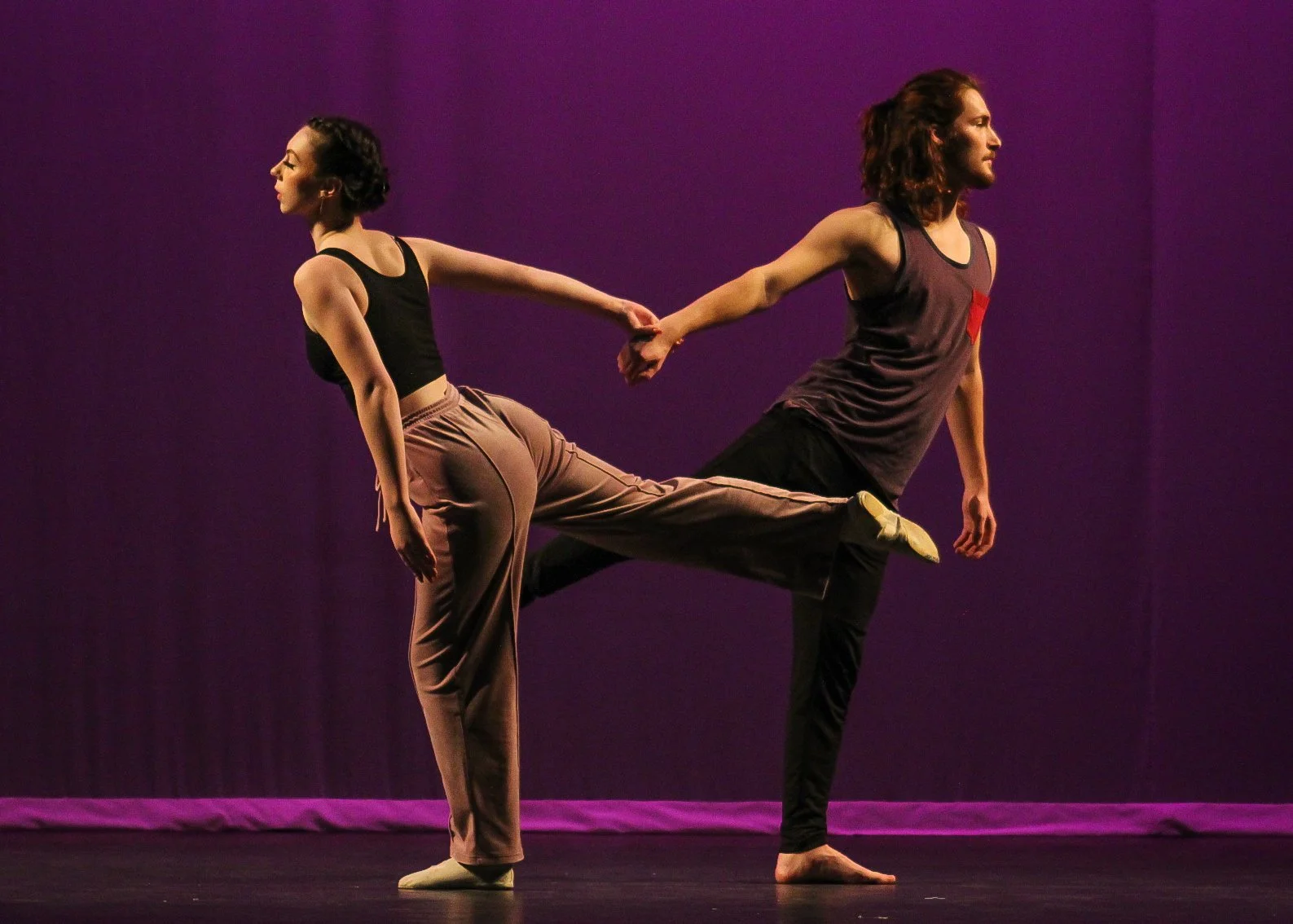 Two performers, a man and a woman, dance on stage against a purple background. The woman is dressed in a black top and beige pants, leaning forward with her eyes closed. The man has long, curly hair and is wearing a sleeveless shirt and black pants. 