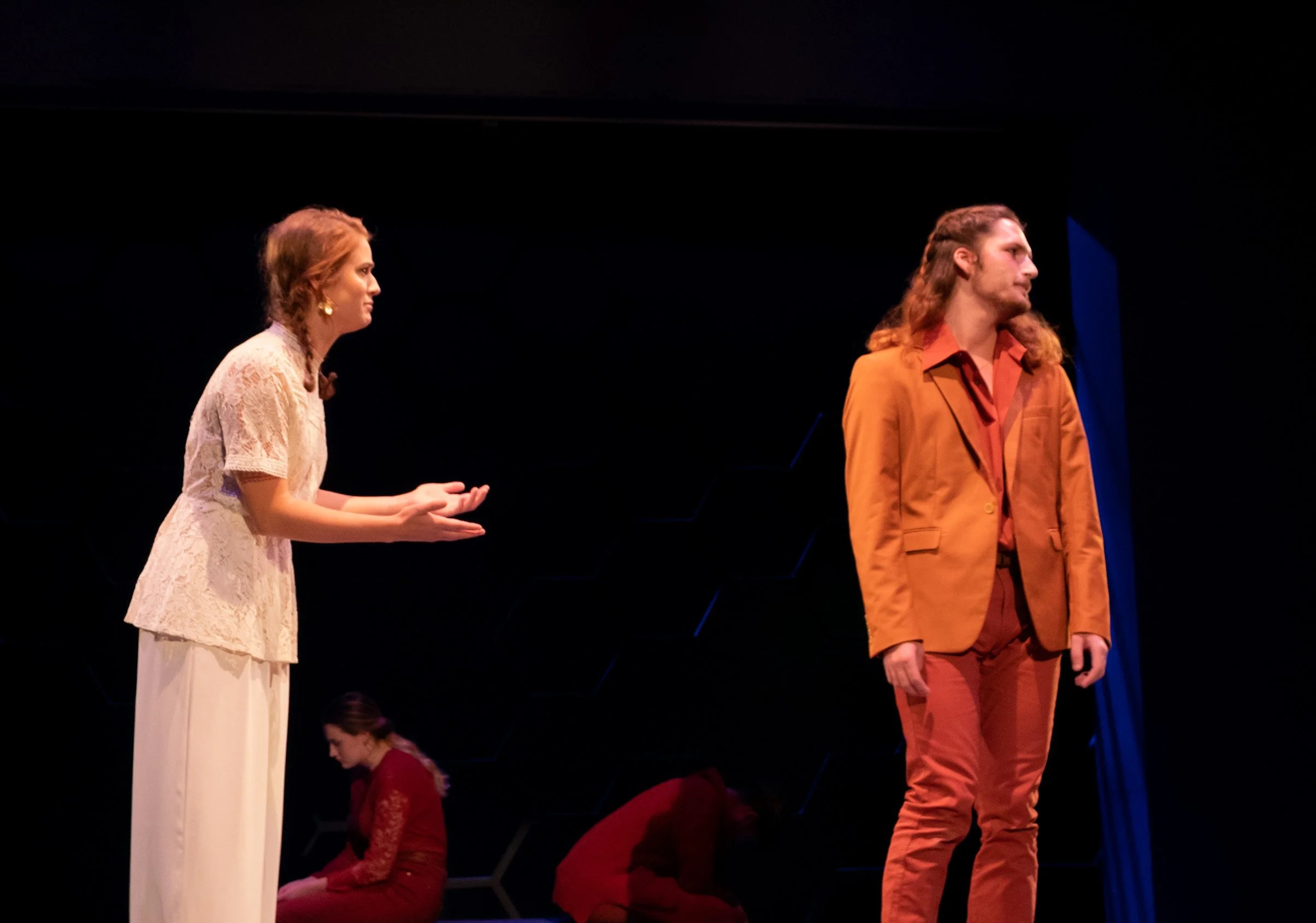 Four actors on stage during a theater performance. Two actors in the foreground, woman in a white lace top and long skirt speaking to man in a brown suit, and two actors in the background sitting, woman in red dress and man in red shirt, with a dark 