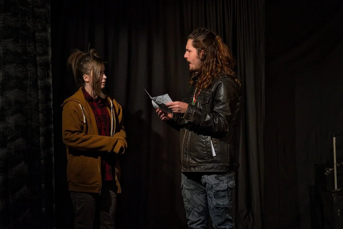Two young adults, a woman and a man, are standing and facing each other against a black curtain background. The woman is wearing a brown hoodie over a red plaid shirt, and the man is wearing a black leather jacket, holding a piece of paper or script,