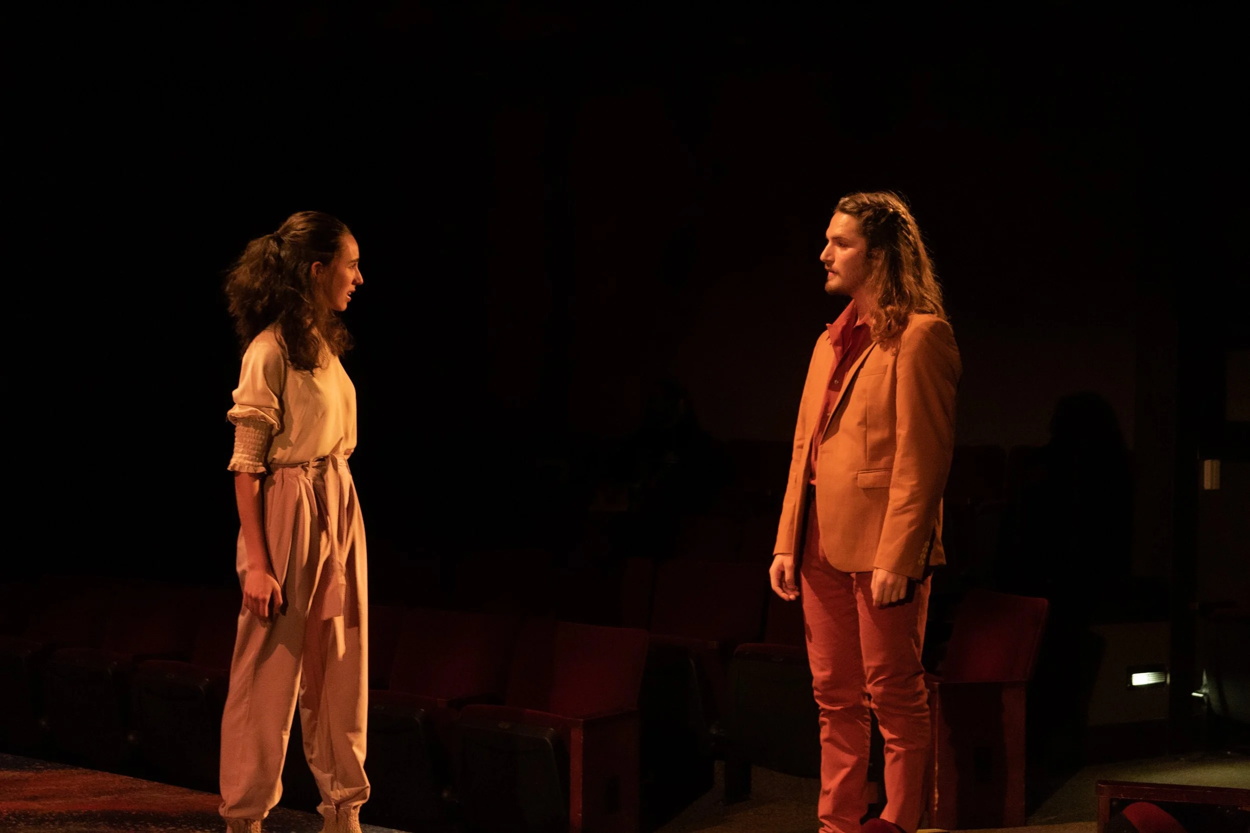 Two people, a woman and a man, stand in a dark room facing each other with a theatrical setting. The woman wears beige pants and a loose beige top, while the man wears a tan suit with a reddish-brown shirt. They are looking at each other with serious