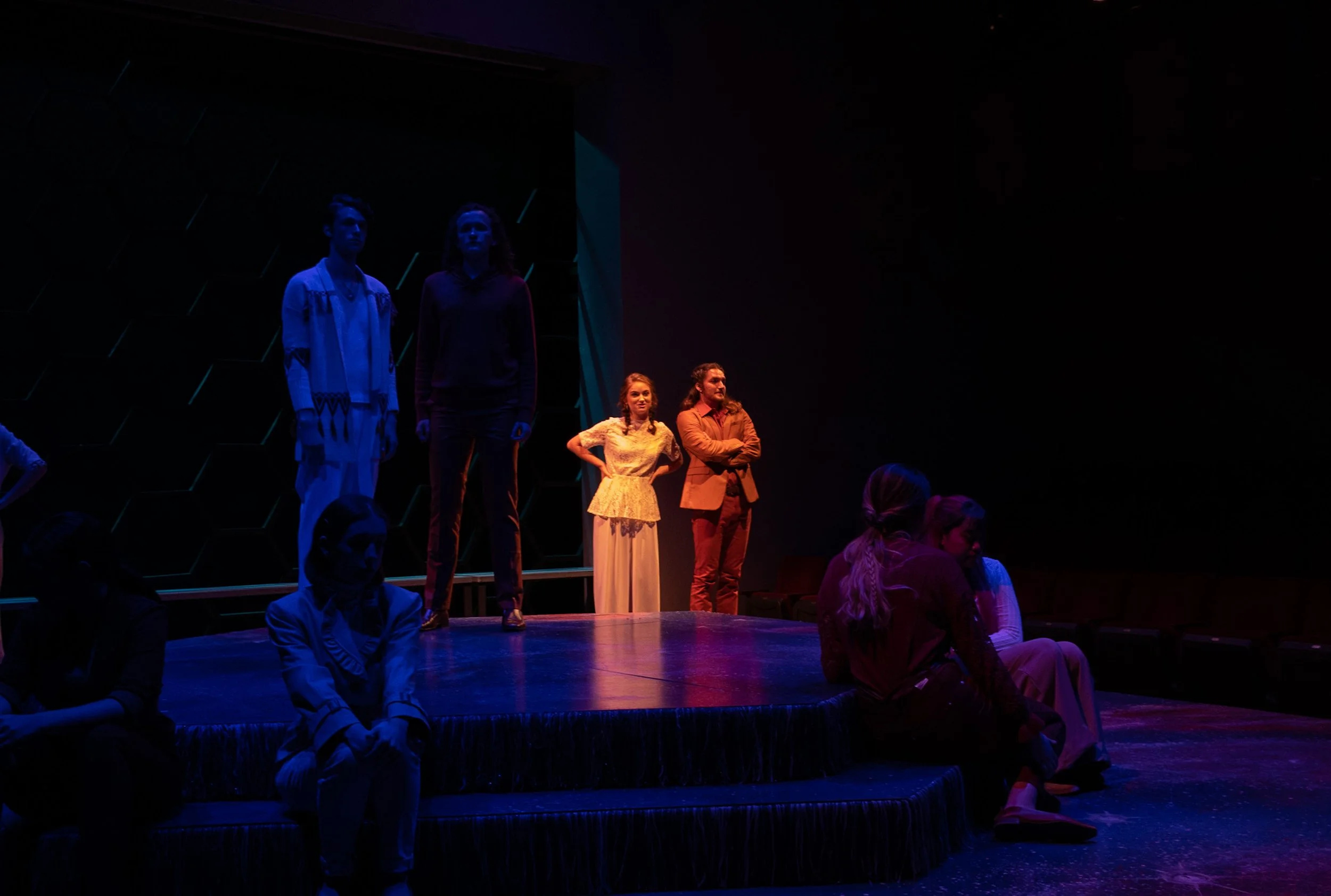 People on a stage in a theater, lit with colorful lights, with some sitting and some standing