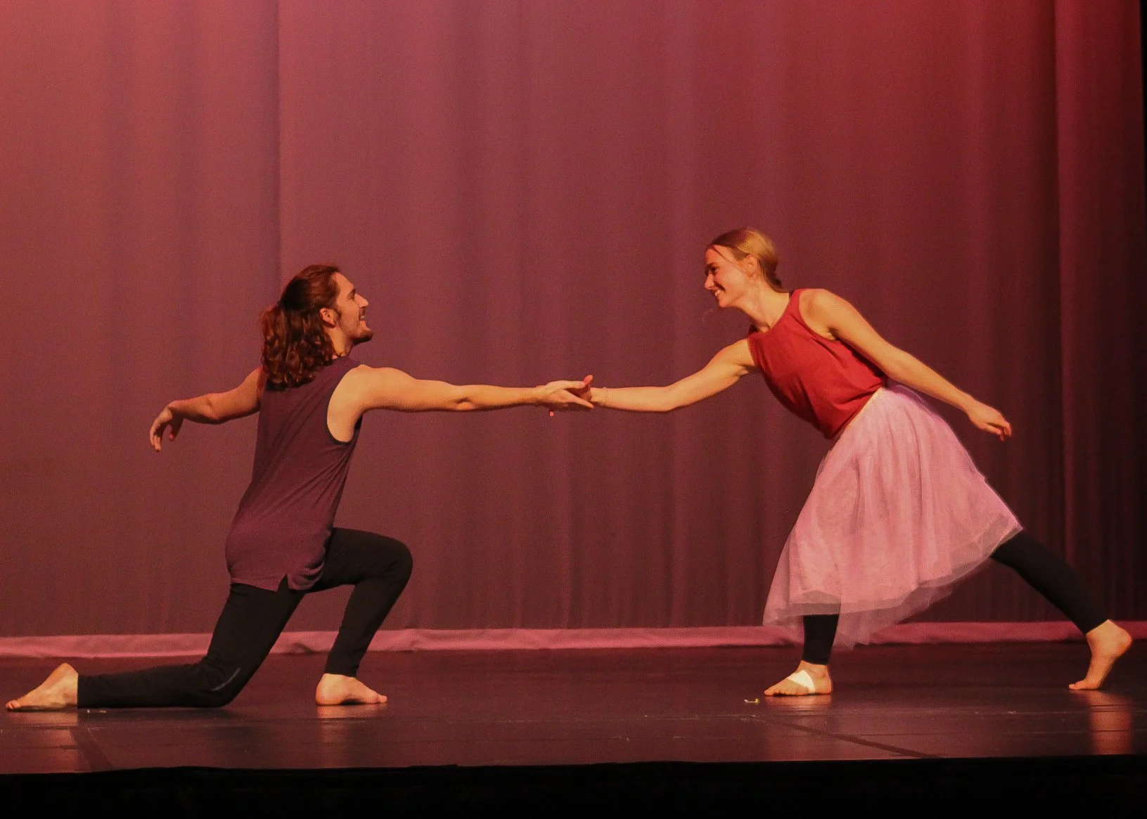 A man and woman dancing on stage, reaching out and holding hands, with pink stage curtains in the background.