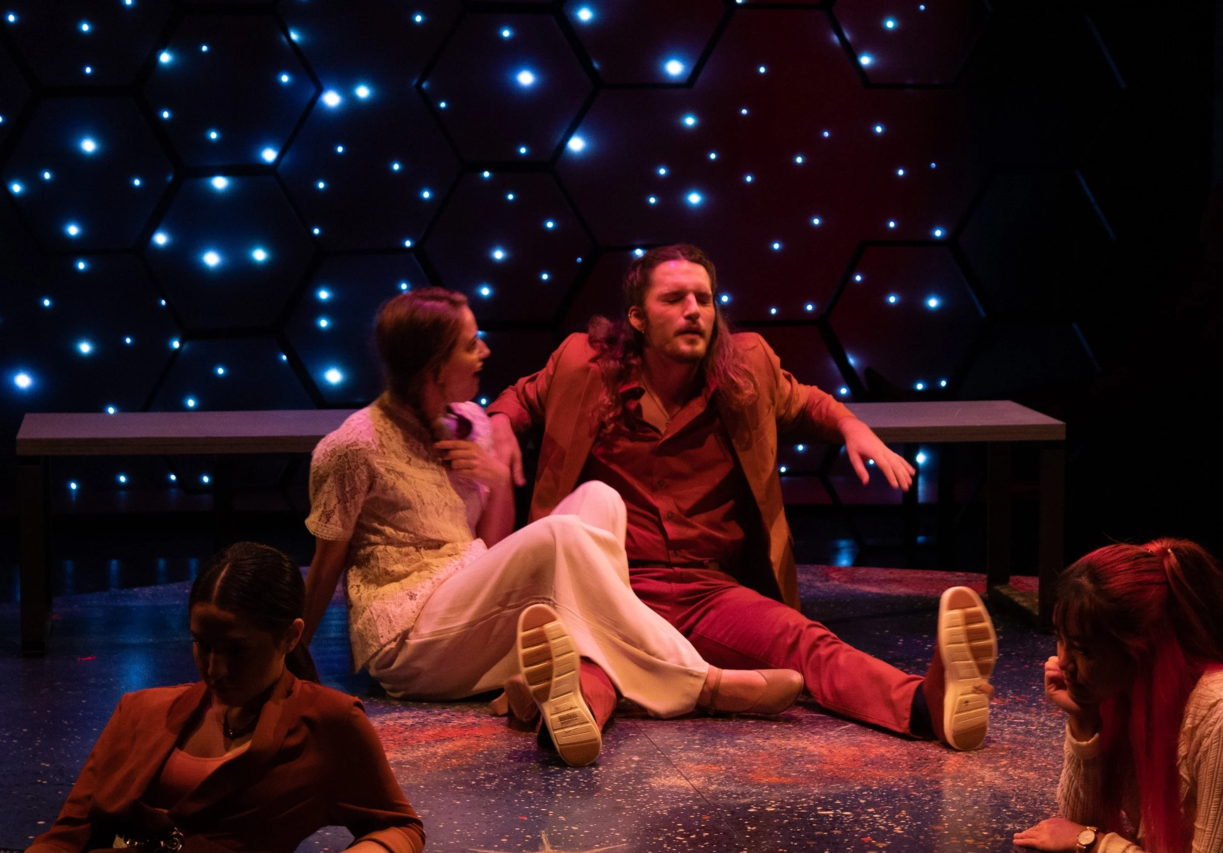 Actors performing on stage with a dark hexagon-patterned backdrop illuminated by small blue lights. One actor with long hair and a brown jacket is sitting on the floor with legs outstretched, eyes closed. A woman in a cream lace top sits close, leani