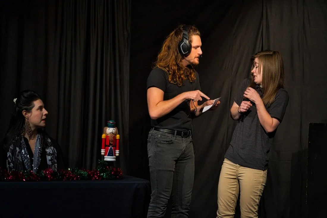 Three people involved in a rehearsal or performance, with one seated at a table with a decorated nutcracker figure, and two others standing and talking, one wearing headphones.