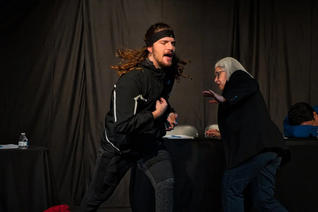 A man with long hair, wearing a black jacket and headband, appears to be upset and is gesturing passionately. An older woman with gray hair and glasses, dressed in black, seems to be scolding or speaking to him sternly. There are tables with papers a