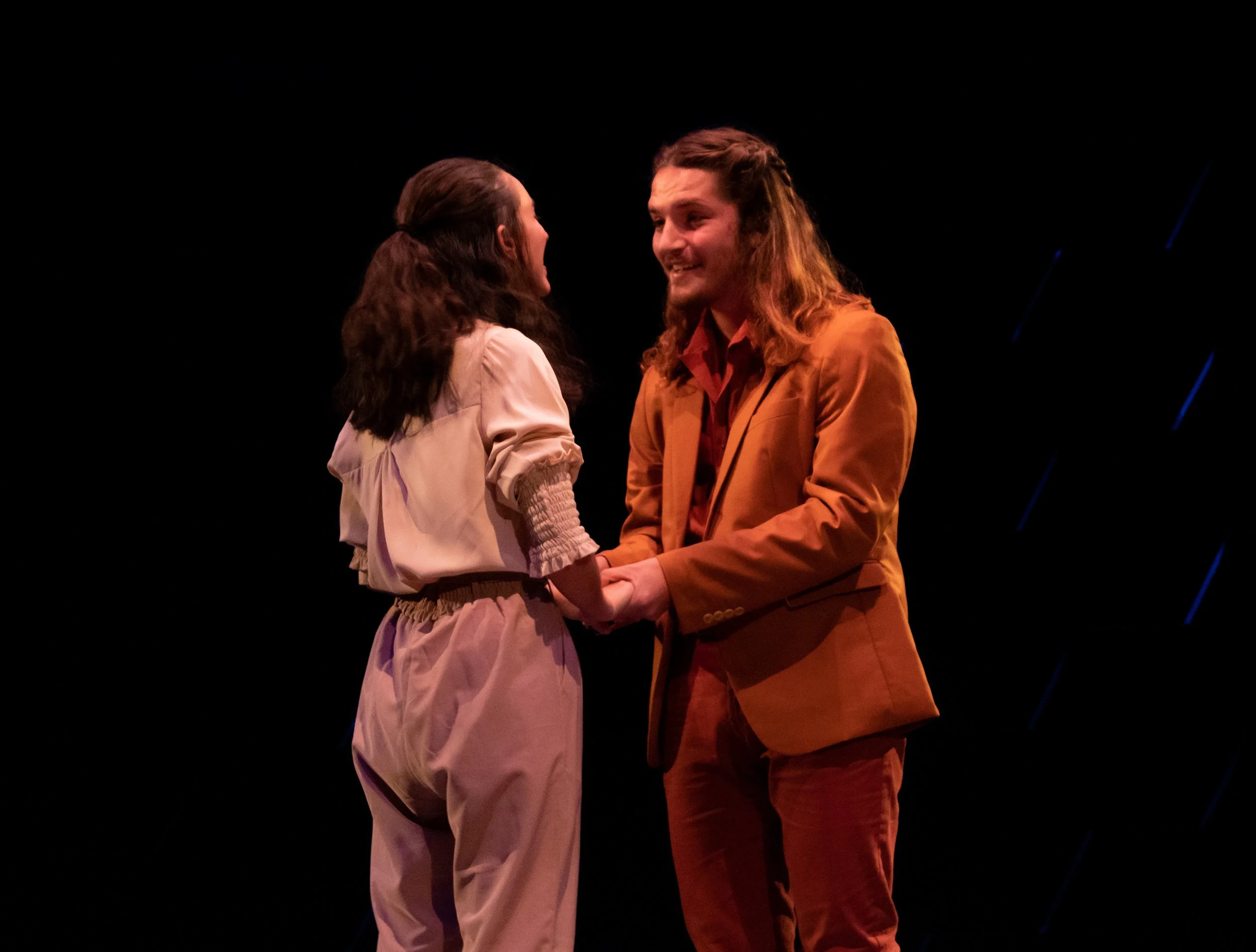 A man and woman holding hands, smiling and looking at each other, on a stage with a black background.