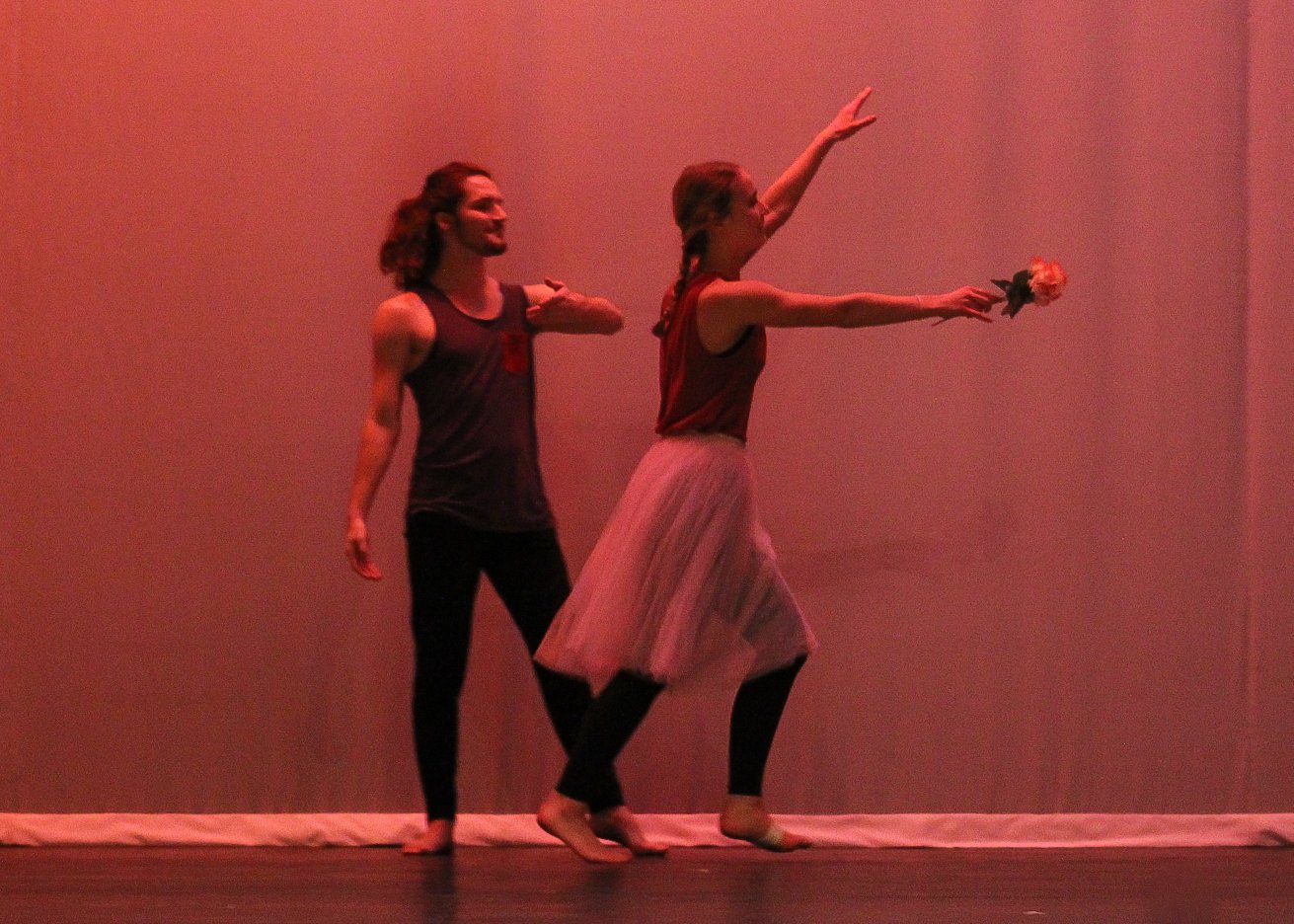 Two performing dancers on stage with pink lighting; one male and one female, with the female holding a flower.