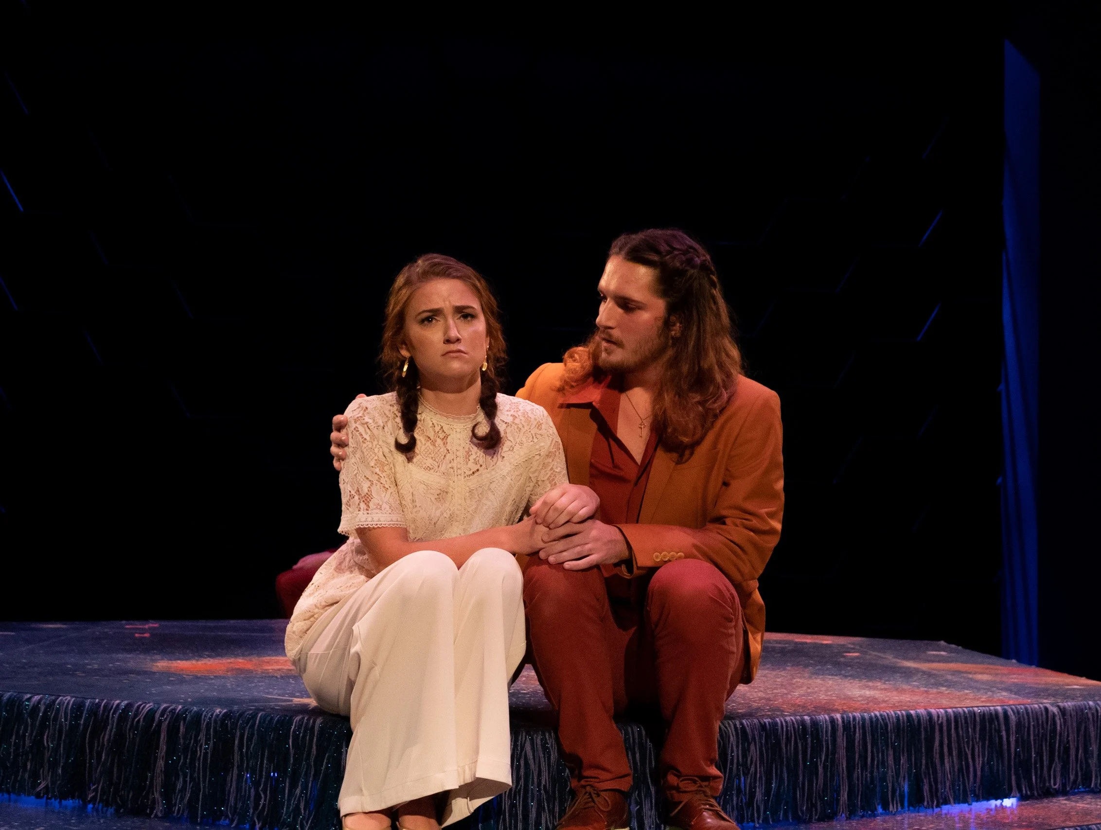A man and woman sitting together on a stage, holding hands, with serious expressions. The woman wears a cream lace top and white pants, the man wears a rust-colored suit. The stage has a dark background with a blue curtain on the right.