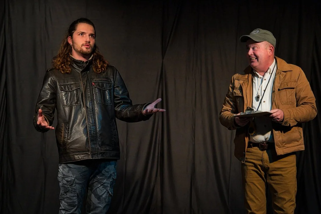 Two men on stage, one with long hair and a leather jacket gesturing, the other wearing a cap and holding a clipboard, smiling. Black curtain backdrop.