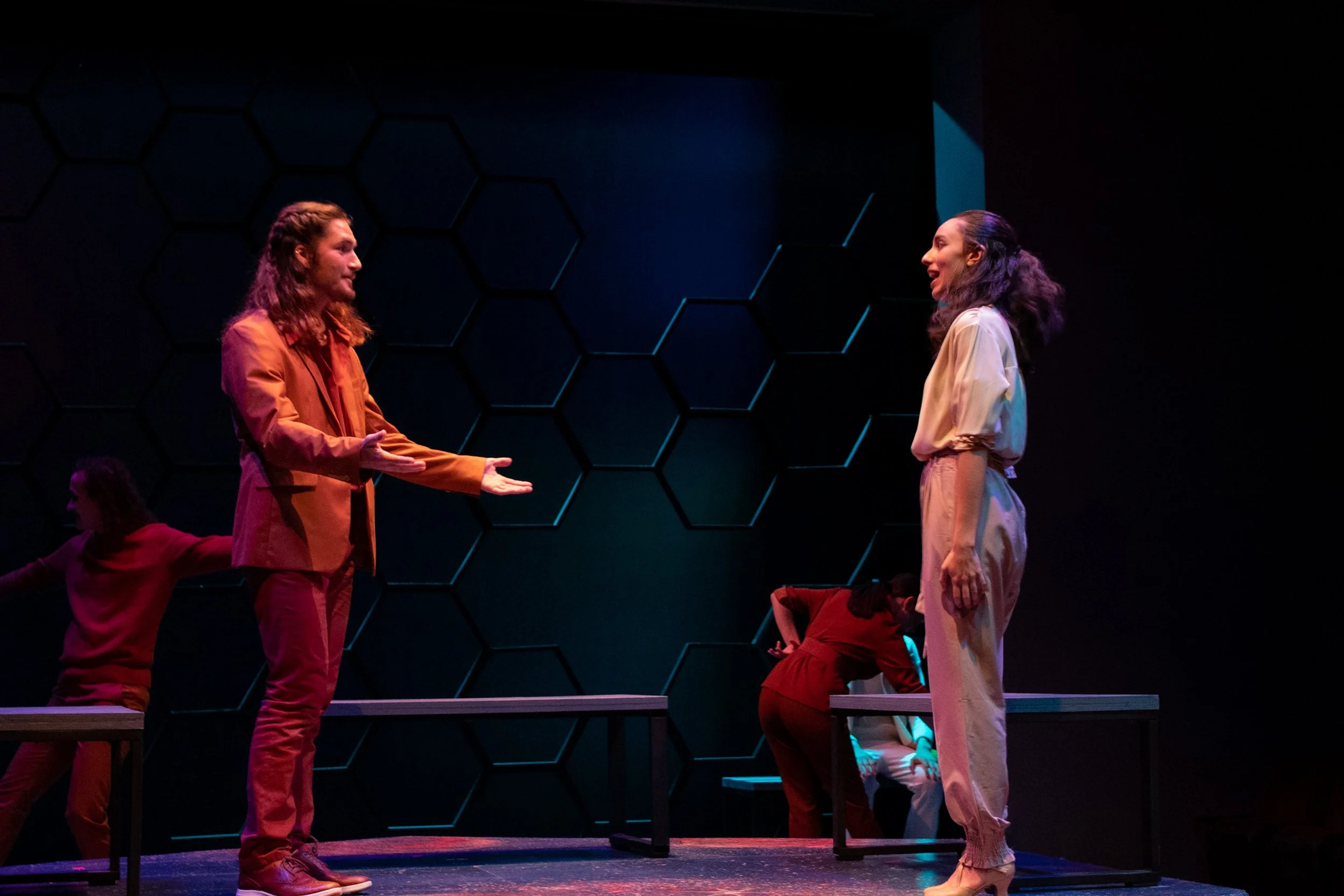 Two people, a man and a woman, are standing on a stage facing each other, engaging in a conversation. The man on the left has long hair, is wearing a brown suit, and is gesturing with his hands. The woman on the right has dark hair tied back, is dres