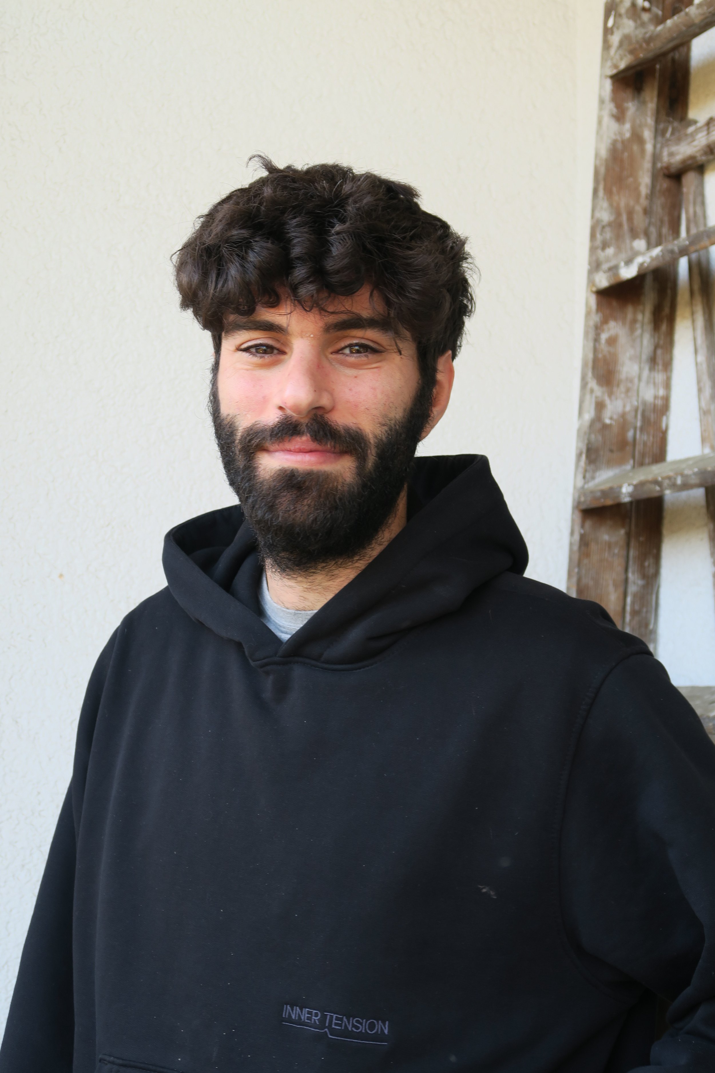 Portrait of a young man with dark curly hair and a beard, wearing a black hoodie, standing outdoors against a light-colored wall with a wooden ladder in the background.