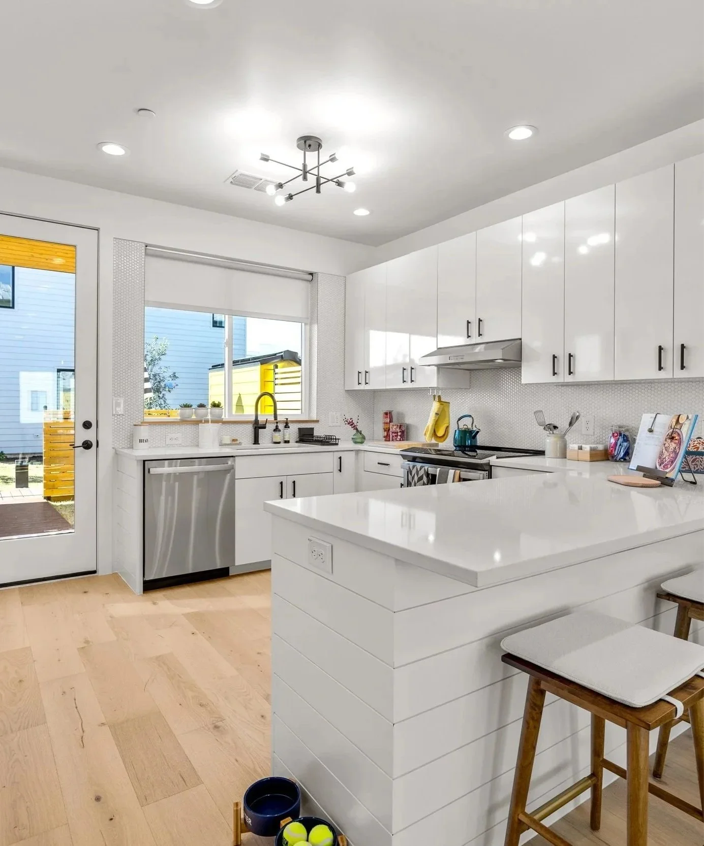 Modern white kitchen with cabinets, stainless steel appliances, and a wooden floor. Via a window and a glass door, outdoor scenery with a yellow shed and neighboring houses can be seen. The kitchen includes a small island with two wooden chairs, a black and white towel hanging from the stove, and various small items and decorations on the counters.