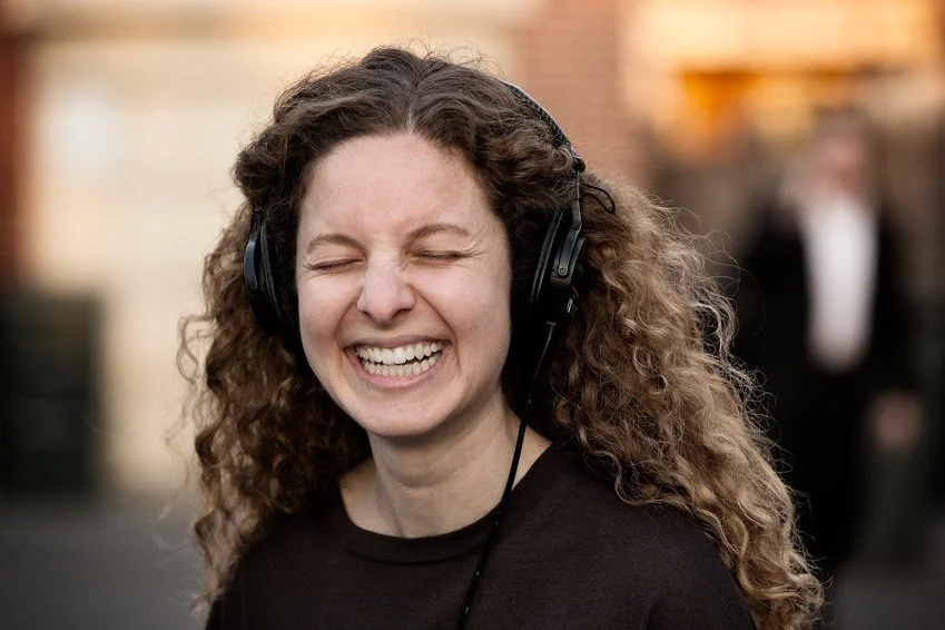 A white woman with long, curly, light brown hair wears headphones and is lighting with her eyes closed.