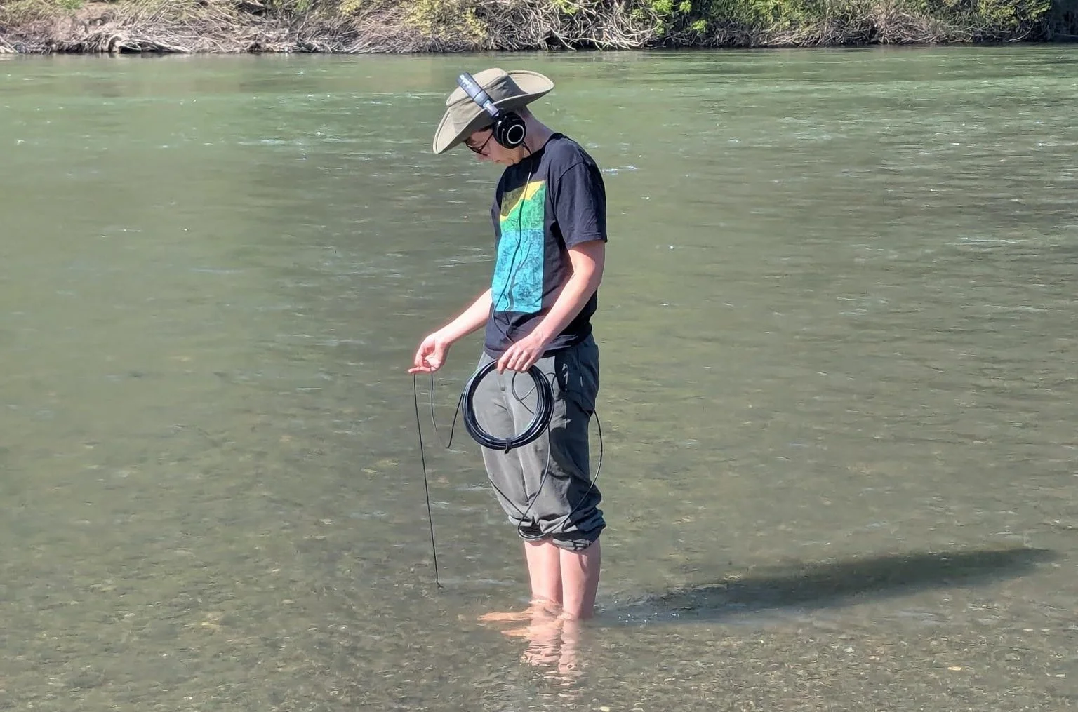 A person with headphones over their booney hat, stands with their pants rolled up to the knee and their feet in a body of water, holding a cable that extends into the water.