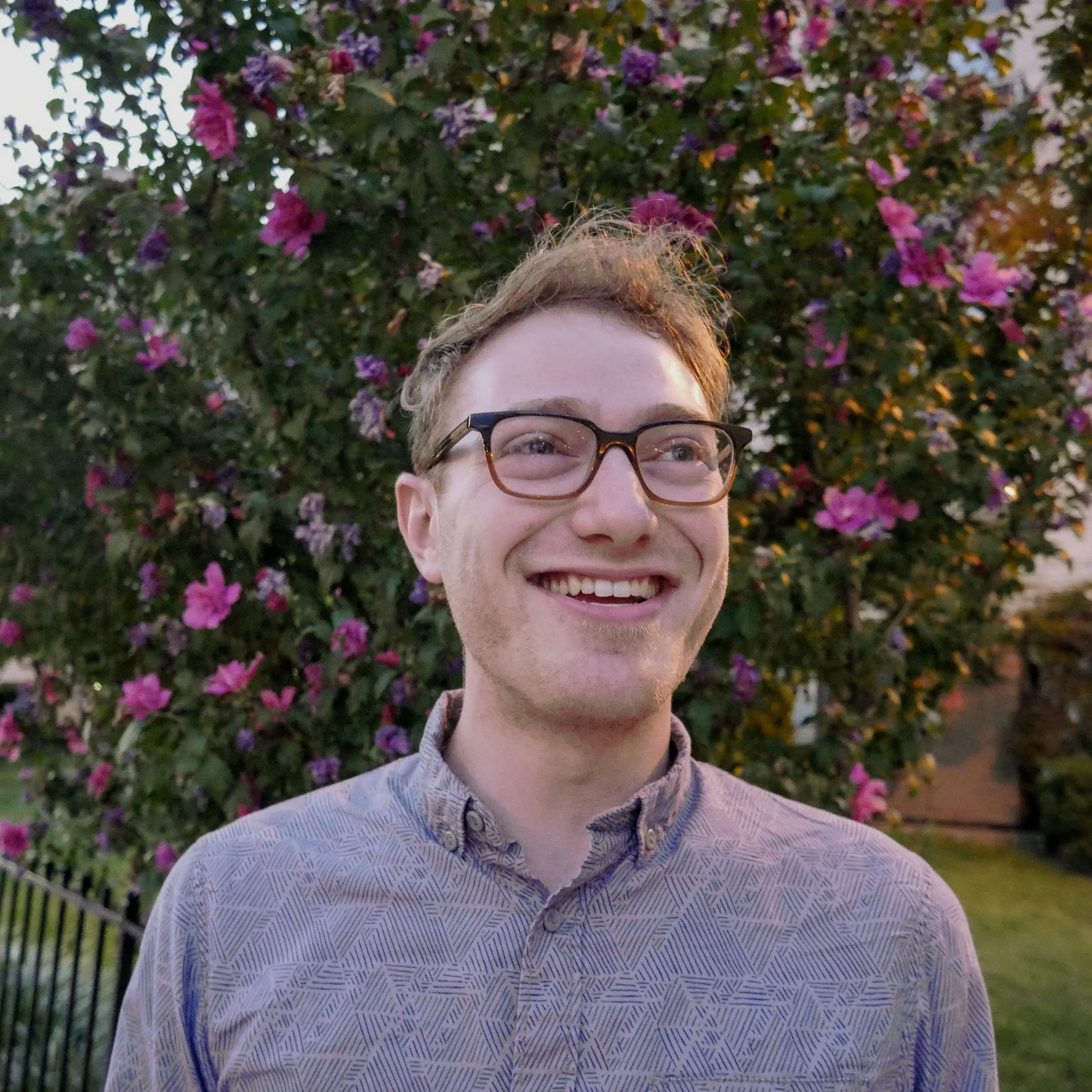 A white male with a light purple shift and dark rimmed glasses smiles with a flowering tree behind him